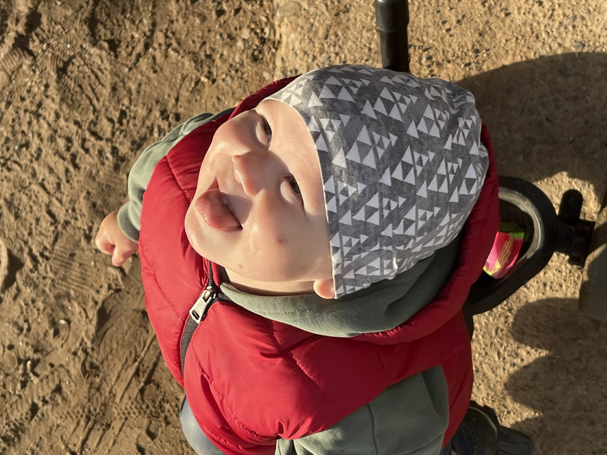 An overhead, close-up shot features a young child, likely a toddler, looking directly upwards with a playful expression, their tongue slightly extended. The child is wearing a grey beanie with a white geometric triangle pattern, a red quilted puffer vest, and a sage green long-sleeved top or hoodie underneath. The bright, warm sunlight casts distinct shadows on the sandy, dusty ground, suggesting it is daytime with clear weather, possibly morning or late afternoon. Footprints are visible in the textured ground. To the right, parts of a black metal bar and a wheel, possibly from a stroller, tricycle, or similar child's equipment, are visible. The setting appears to be an outdoor, unpaved area in Фоки, Russia. There is no visible text in the image.
