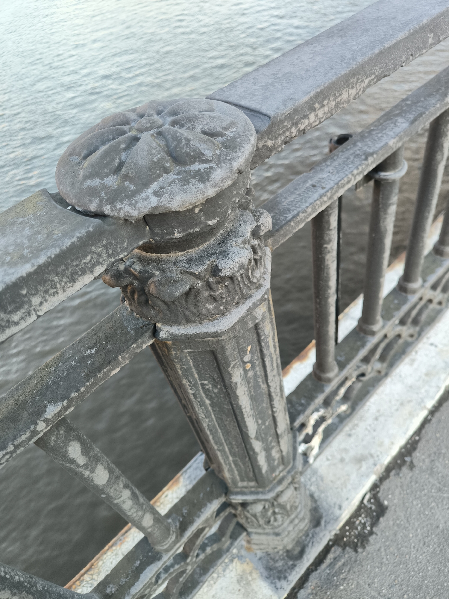 The image shows a close-up, eye-level view of a weathered metal railing on a bridge. The railing is dark gray with visible signs of age and wear, including patches of white residue, possibly frost or salt, and some rust. The railing features decorative elements, including a floral-shaped cap on a post and ornate carvings on the post itself. Behind the railing, a body of water, likely a river, is visible, its surface rippled. The water appears to be a muted gray-blue. The bridge's edge, made of concrete or stone, is visible on the right side of the image, showing similar signs of weathering. The overall impression is one of an older, possibly historic, piece of urban infrastructure. The scene is devoid of people or any signs of recent activity. The context of Moscow, Russia, is not directly evident from the visual elements alone, but the description suggests a potentially cold climate given the presence of white residue that could be frost.