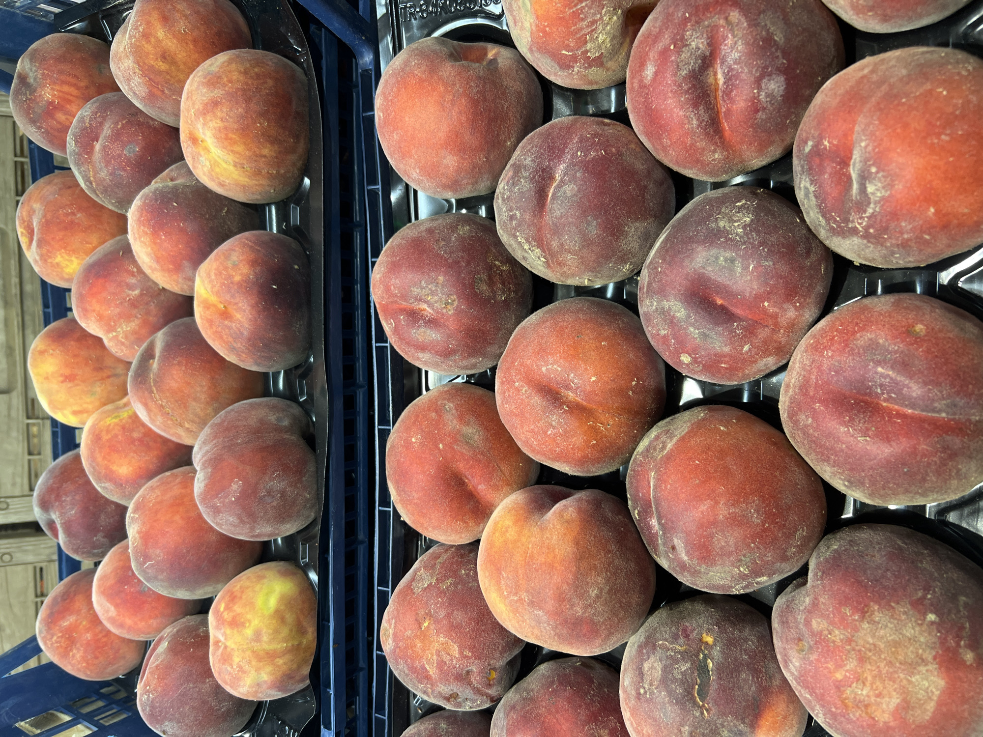 The image shows two plastic crates filled with ripe peaches. The crates are stacked on top of each other, with the top crate positioned in the foreground and the bottom crate behind it. The peaches in both crates are predominantly red and orange, with some yellow undertones. They appear to be fresh and in good condition. The background is blurred but suggests an indoor environment, possibly a market or a storage area. There are no people or specific landmarks visible to indicate a location in Fontanka, Ukraine.