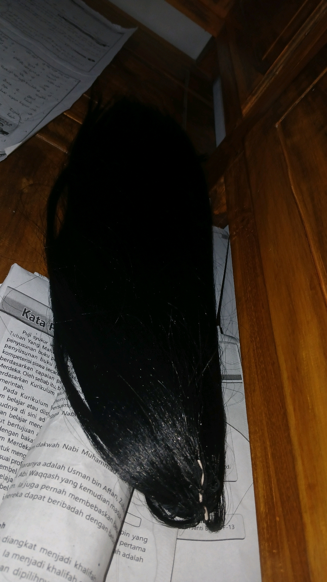 The image shows a close-up of a bundle of black hair on top of open books. The books appear to be school textbooks, with Indonesian text visible on the pages. The hair is long and straight, with some strands appearing to be synthetic or attached to something. The background is a wooden surface, possibly a table or desk. The lighting is dim, creating shadows and highlights on the hair and books. The overall scene suggests an indoor setting. There is no discernible location within the city of Sampang, Indonesia.