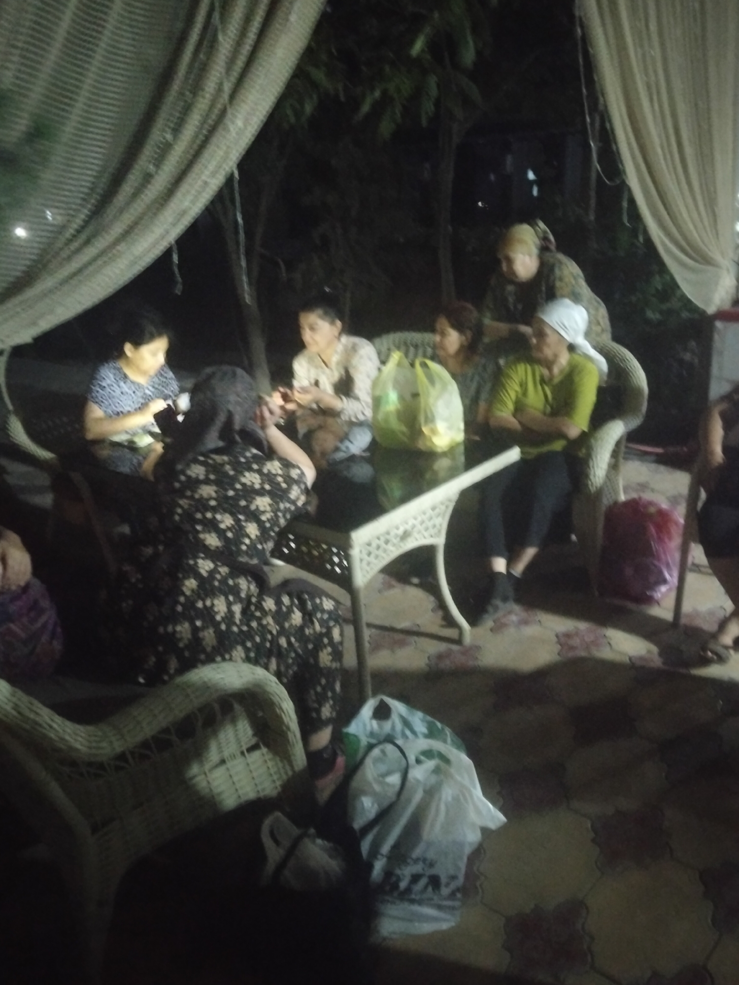 The image depicts an outdoor scene at night, possibly in Kagan, Uzbekistan, with a group of people gathered around a table. Several women are seated, some wearing headscarves. One woman in a green shirt is seated in a wicker chair with her arms crossed. Another woman in a black and white floral dress is seated facing away from the camera. A table with a decorative white base is placed in the center, with shopping bags on and near it. The ground is paved with patterned tiles. Curtains hang from above, and trees are visible in the background. The lighting suggests artificial illumination.