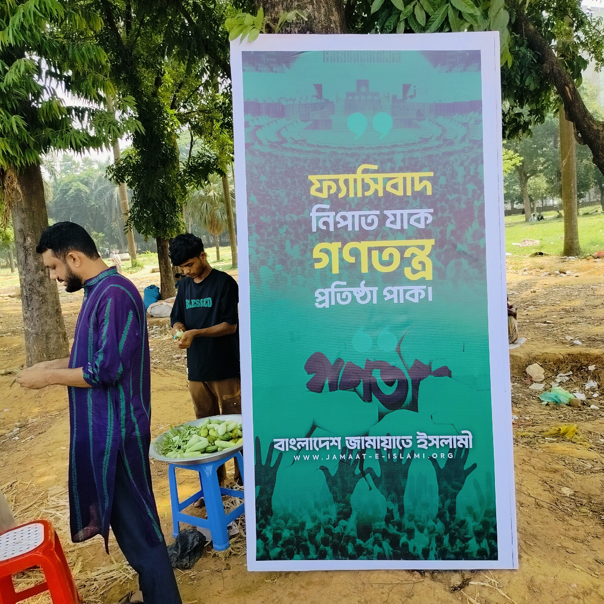 The image depicts an outdoor scene in what appears to be a park or public space in Dhaka, Bangladesh. Two men are visible; one is wearing a purple and green striped shirt and appears to be preparing food. A blue stool holds a tray of vegetables, possibly cucumbers. The second man, wearing a black "BLESSED" t-shirt, is peeling or preparing something. Behind them, a large poster for Bangladesh Jamaat-e-Islami is prominently displayed. The poster includes Bengali script, and the website address www.jamaat-e-islami.org. The background includes trees, grass, and other people in the distance. The ground is dirt and appears to be somewhat littered.