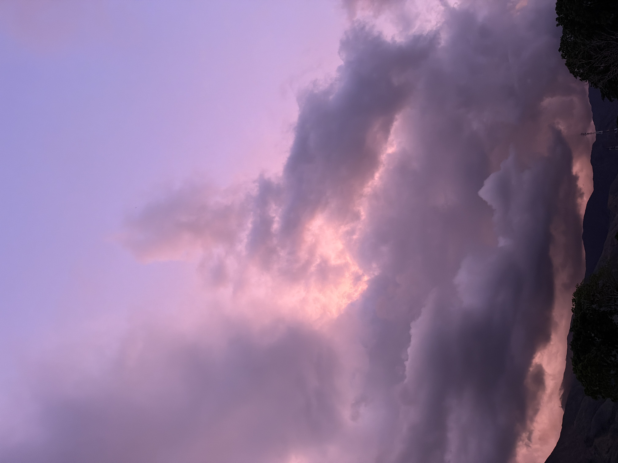 The image captures a dramatic sky over the foothills of El Cambur, Venezuela. A vast expanse of clouds dominates the frame, colored in shades of deep purple, lavender, and pink. Some clouds are dense and dark, while others are illuminated from within by a soft, warm light, suggesting either sunset or sunrise. The upper portion of the sky transitions to a lighter lavender hue.
Along the bottom edge of the image, the silhouettes of dark green trees and the undulating lines of mountains are visible. The land appears mountainous and rugged. A slender communication tower is faintly visible in the distance, rising above the treeline. The overall scene evokes a sense of natural beauty and atmospheric drama. There are no people or specific actions depicted.