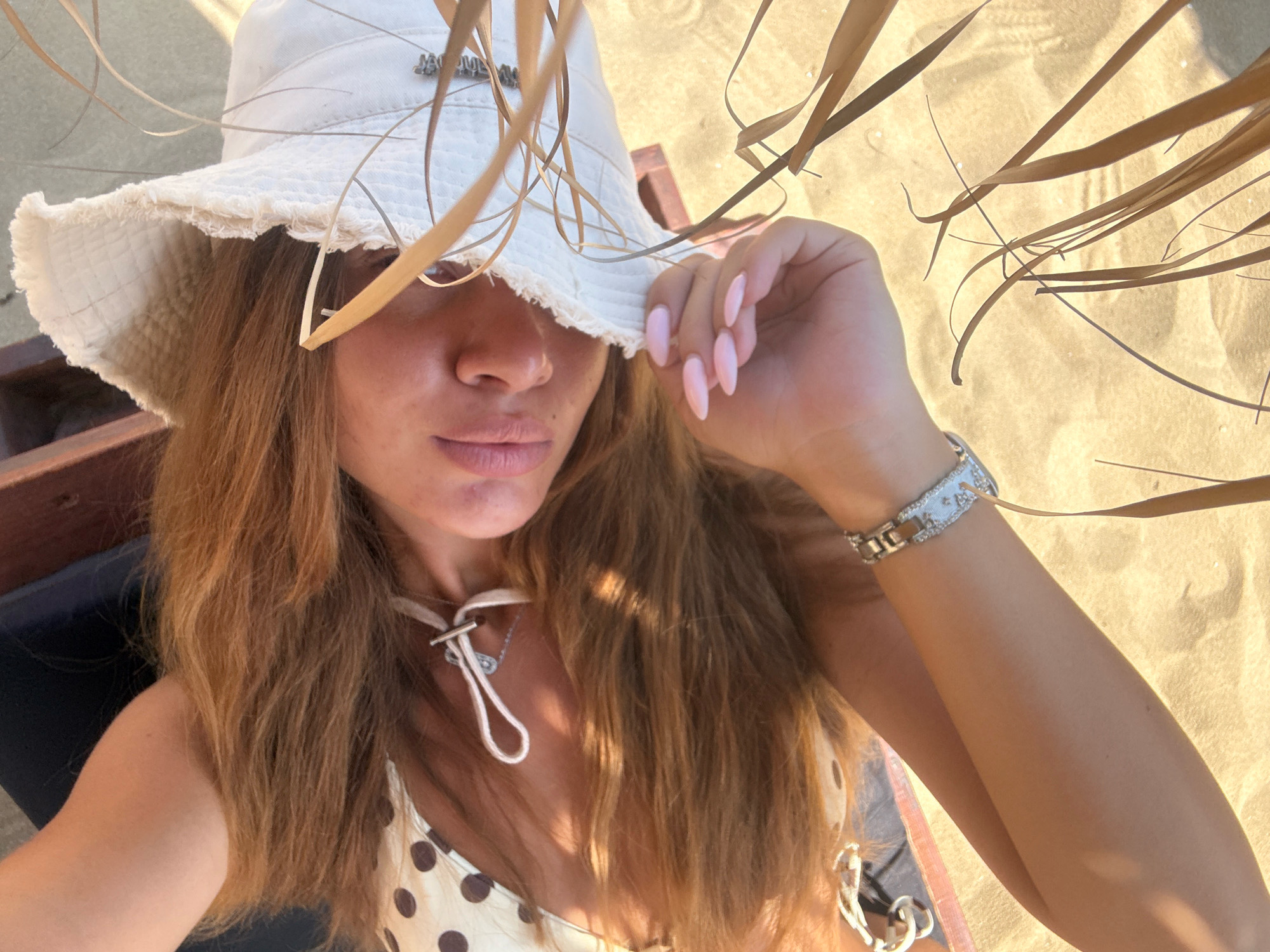 A woman in a white bucket hat and a polka dot bikini is sitting on a beach chair on the sand in Nea Peramos, Greece. She is holding her hat and looking at the camera. There are dried palm fronds in the foreground.