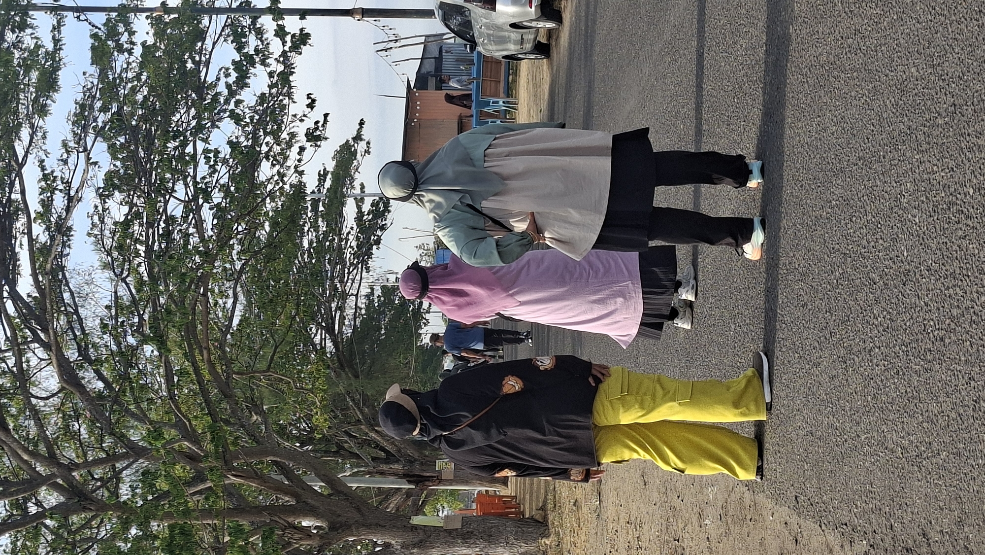 In Lhokseumawe, Indonesia, three women, viewed from behind and walking on a paved road, are the primary subjects. The woman on the left wears a black jacket with orange patterns, a black cap with a tan brim, and bright yellow cargo pants. The middle woman is dressed in a long pink hijab and skirt, also wearing a cap. The woman on the right is clad in a light green and beige hijab and an outfit with a long, flowing top over dark pants. They are walking in the same direction along the road, which appears to be a pedestrian walkway or shoulder, with a dirt area to the left and trees lining the side. In the background, a silver SUV is parked on the right side of the road, and beyond that, there are buildings, some with awnings, and a clear sky. The overall scene suggests a daytime outdoor activity, possibly in a park or along a coastal area given the presence of trees and open space.