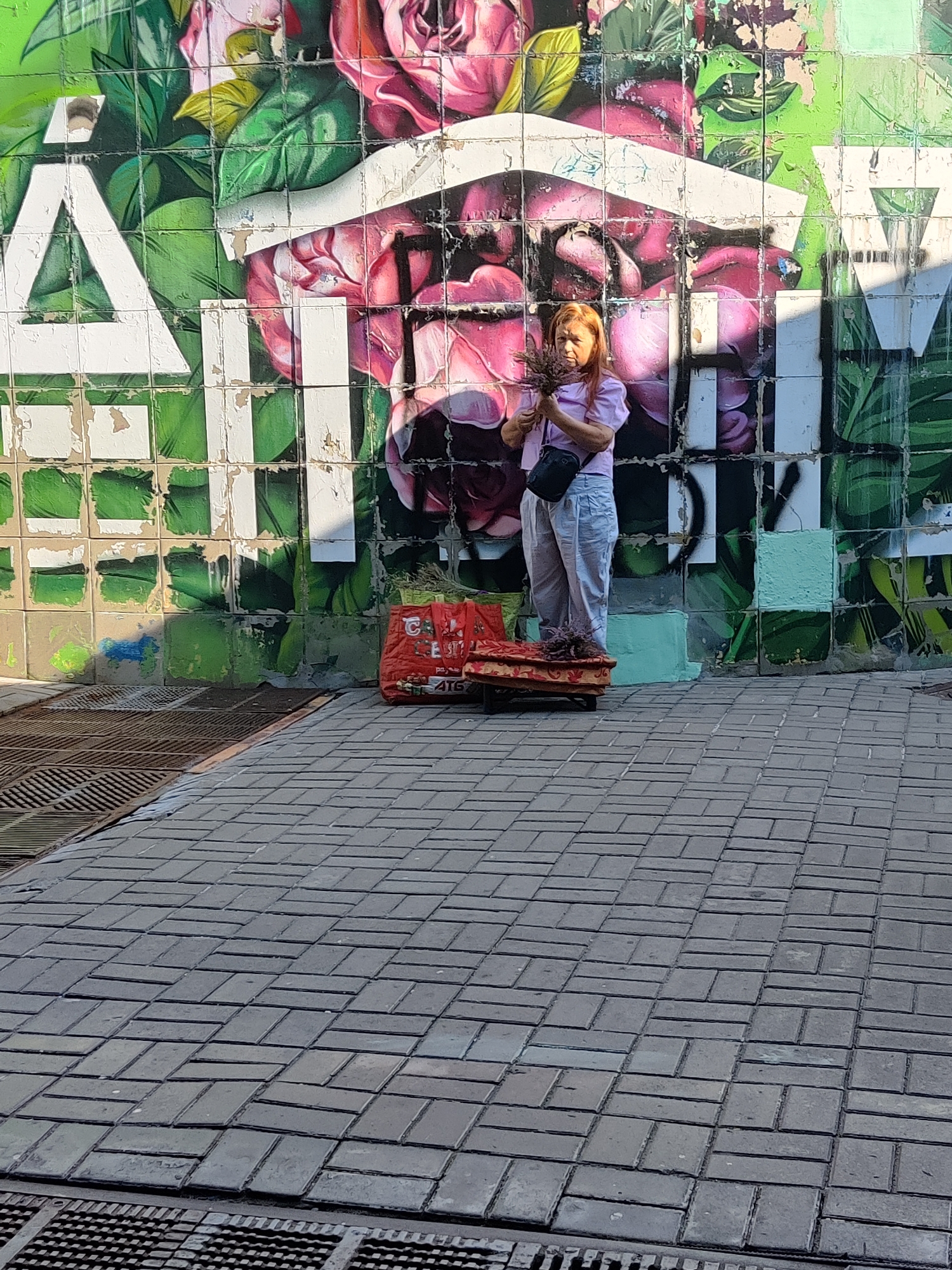 A woman with red hair stands on a raised, fabric-covered platform in front of a graffiti-covered wall in Kyiv, Ukraine. The graffiti depicts large, stylized pink roses and green leaves. The woman is wearing a light pink shirt and light-colored pants, and she holds a small bouquet of dried flowers in her hands. She has a black crossbody bag. To her left, on the ground, is a large red bag. The ground in front of the wall is paved with rectangular gray bricks. To the left of the paving, there is a metal grate. The scene appears to be outdoors.