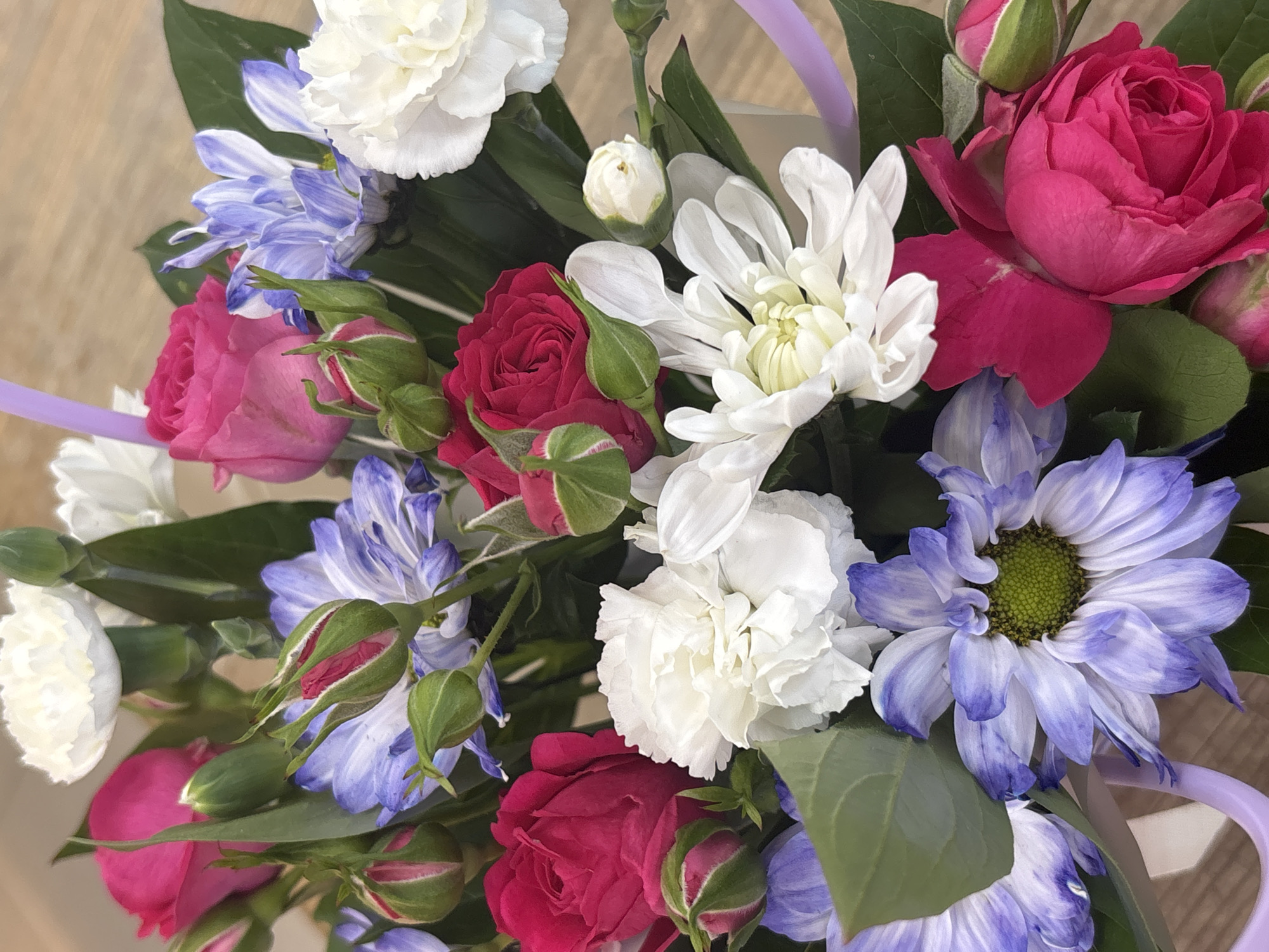 A close-up, overhead shot features a vibrant bouquet of flowers, including pink and red roses, white carnations, and purple and white aster-like flowers. The flowers are arranged tightly together with green leaves and buds interspersed throughout. A pale purple ribbon is visible weaving through the bouquet. The background is a blurred, light brown surface, possibly a table. The lighting is bright and even, highlighting the rich colors and textures of the floral arrangement. There is no discernible connection to Chaikovsky, Russia, nor any people or actions depicted.