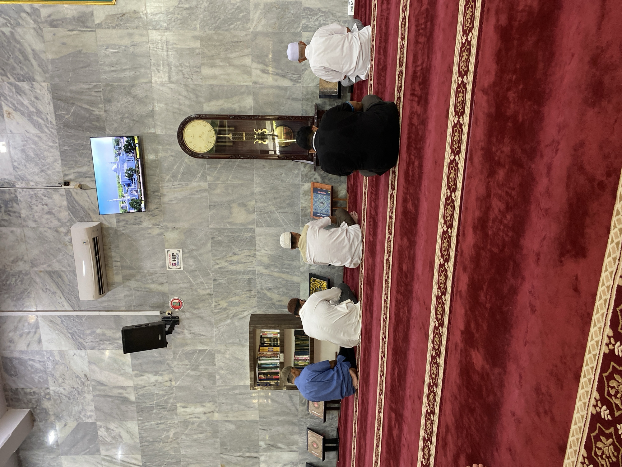 This image depicts the interior of a mosque, likely in Lhokseumawe, Indonesia. Five men are seated on a plush red carpet, facing the same direction, indicating they are engaged in prayer or a religious gathering. The wall behind them is adorned with grey marble tiles.
To the left, a man in a blue shirt is seated, with a bookshelf filled with religious texts behind him. Next to him, two men in white attire are seated. The central man is facing away from the camera, while the man to his right is also facing away, with a book open on a stand in front of him. Further to the right, a man in a black shirt is seated. On the far right, a man in white attire, wearing a white cap, is also seated and facing away.
Above the men, an air conditioning unit is mounted on the wall. To the right of the AC unit, a black speaker is mounted on the wall. A small sign with the letters "HP" and Arabic script is visible. A large, ornate grandfather clock stands vertically on the right side of the image, its pendulum visible through a glass casing. A flat-screen television is mounted on the wall above the clock, displaying an image of a white domed mosque. The red carpet features decorative patterns along its length. The lighting suggests an indoor setting, likely during daylight hours.