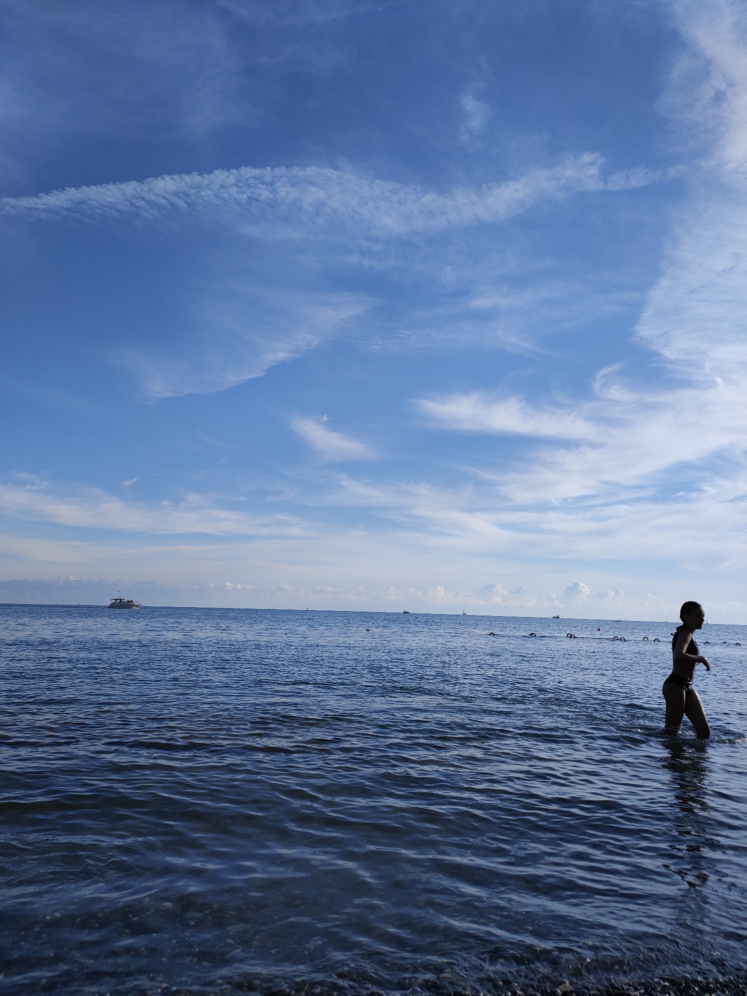 A young woman in a black bikini walks through shallow water at the edge of the sea. She is silhouetted against a bright blue sky filled with wispy white clouds. In the distance, a white boat is visible on the horizon, along with a line of buoys marking a swimming area. The water is calm, with small waves lapping at the shore. The scene appears to be a beach in Sochi, Russia, on a clear day.