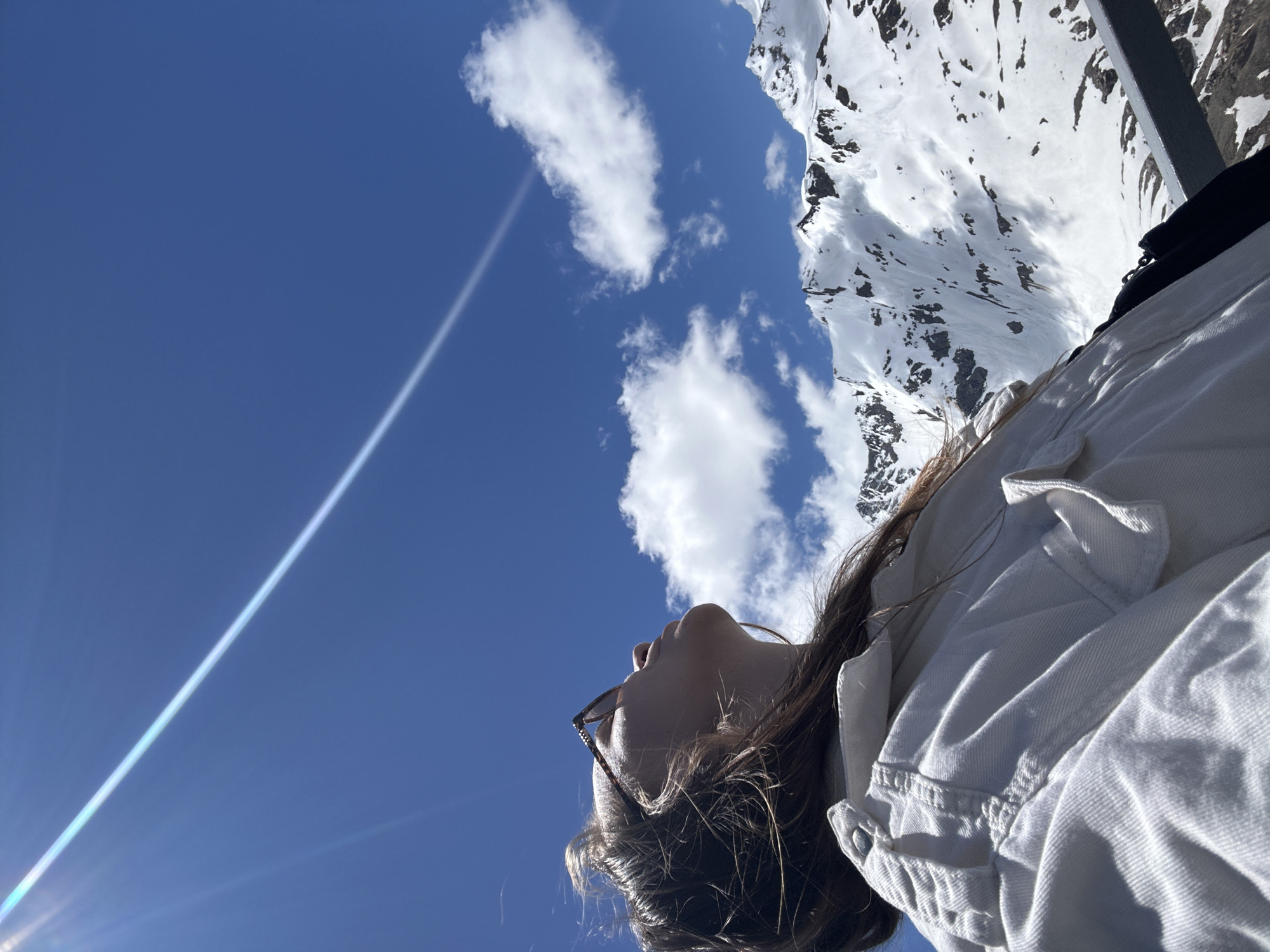 In Terskol, Russia, a young woman with long brown hair, wearing sunglasses and a white jacket, looks up at the sky against a backdrop of snow-capped mountains and a clear blue sky. Sunlight streaks across the sky, creating a lens flare. The woman appears to be standing on an elevated platform, as indicated by a metal railing in the foreground. The surrounding environment is mountainous and appears to be a winter or early spring scene, with patches of snow visible on the rugged terrain.