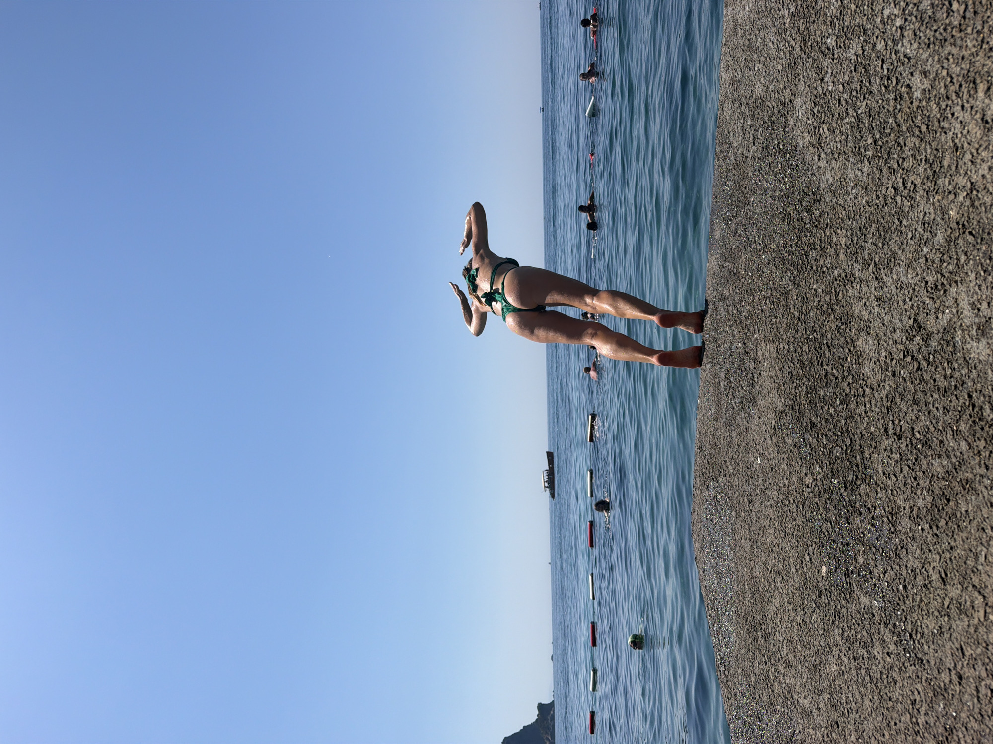 The image depicts a sunny day at the beach in Budva, Montenegro. In the foreground, a woman wearing a green bikini is captured mid-leap, preparing to dive into the blue sea from a concrete pier. Her body is angled downwards, arms raised behind her head, and legs extended. The pier's textured surface is prominent in the lower portion of the frame. In the background, the clear blue sea stretches towards the horizon, with a few buoys marking a swimming area. Several people are visible in the water, some swimming and others on inflatable mats. A small boat navigates the water in the distance, and a mountainous coastline is visible on the left side of the image. The sky is a vibrant, cloudless blue.