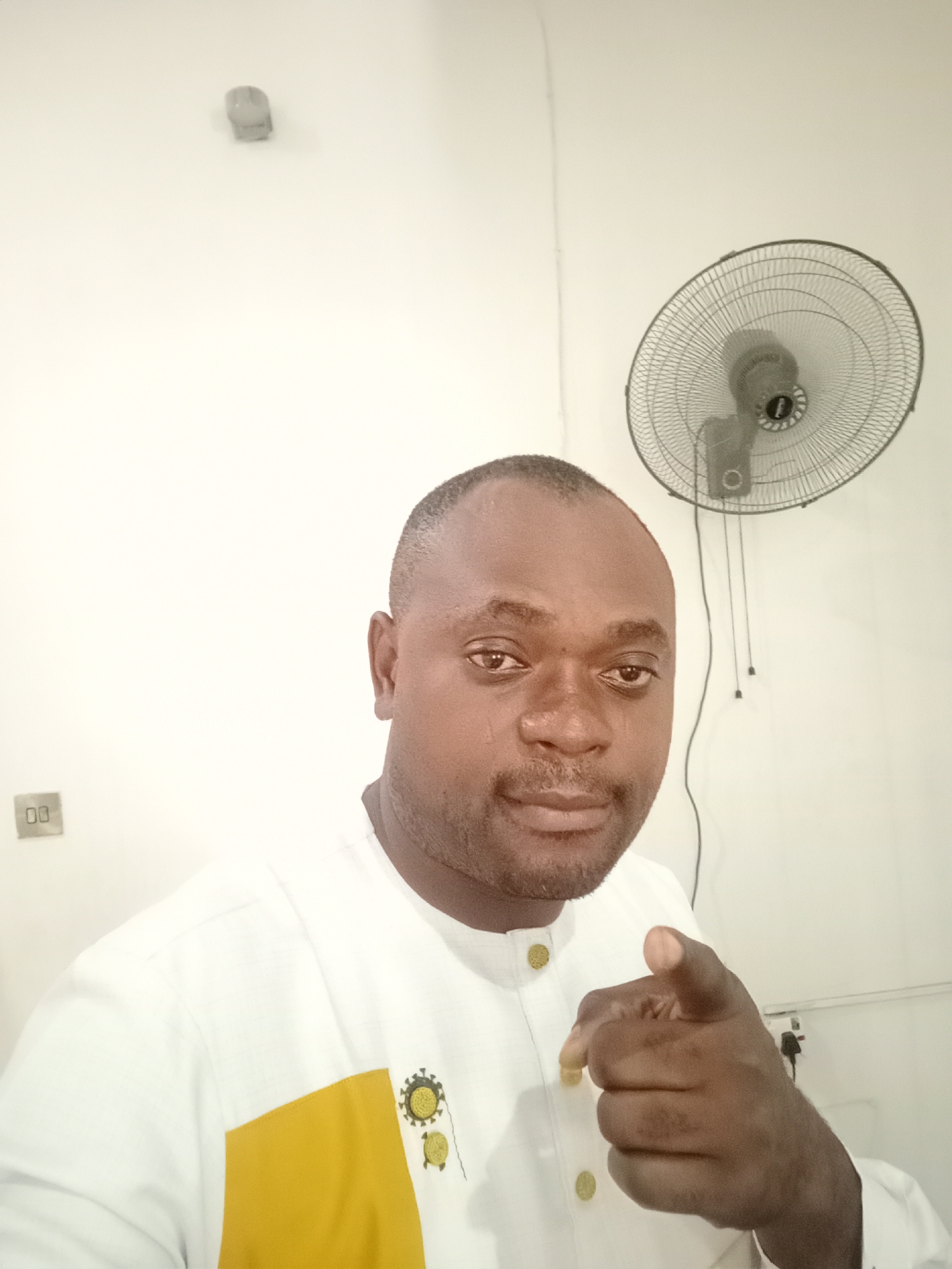 A man is indoors in what appears to be a home or office setting. He is pointing directly at the camera with his right hand. The man is wearing a white shirt with a yellow patch on the left side of his chest, adorned with small gold buttons. He appears to be of African descent.
Behind him is a plain, light-colored wall. Mounted on the wall is an electrical outlet and an oscillating fan. The location is potentially Gwarinpa, Nigeria.