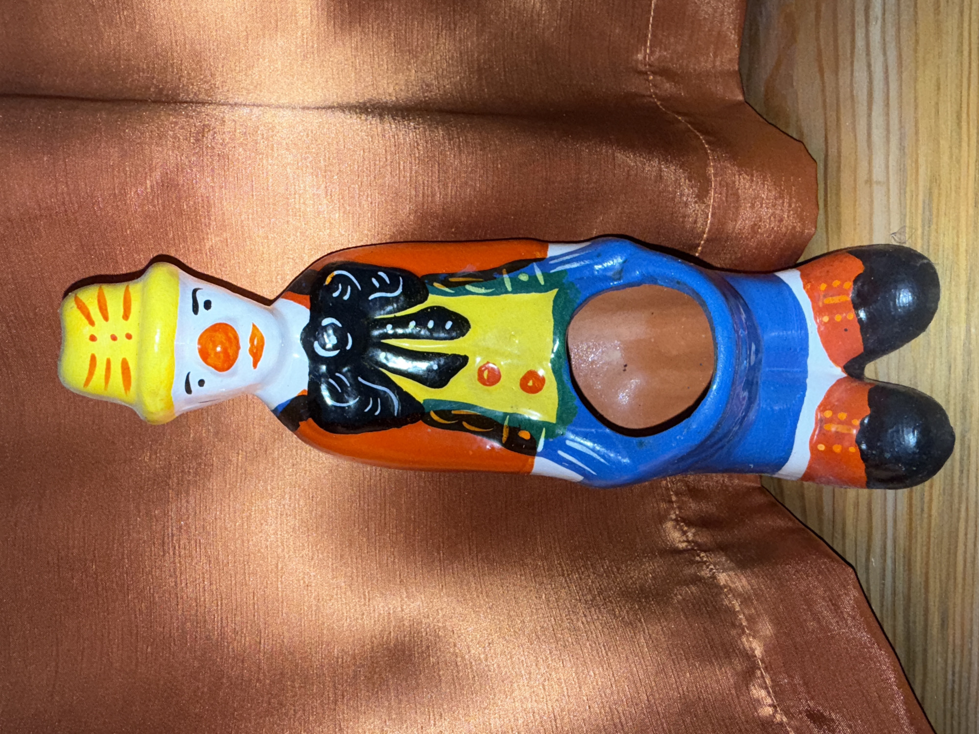A full-body, eye-level, medium shot captures a colorful ceramic clown figurine standing upright. The clown wears a yellow hat with orange stripes, a white face with a red nose and mouth, and black eyebrows. Its outfit consists of an orange top with a black bow tie, a yellow vest with red buttons, and blue pants. The figurine has a hollow opening in its midsection, revealing an inner brown ceramic surface. The clown's feet are black with orange accents. The background is a textured, rust-colored fabric that appears to be a curtain, with folds and shadows visible. The figurine rests on a light-colored wooden surface. The scene is brightly lit, casting minimal shadows. There is no indication of the city of Balaklavskyi Okruh, Ukraine.