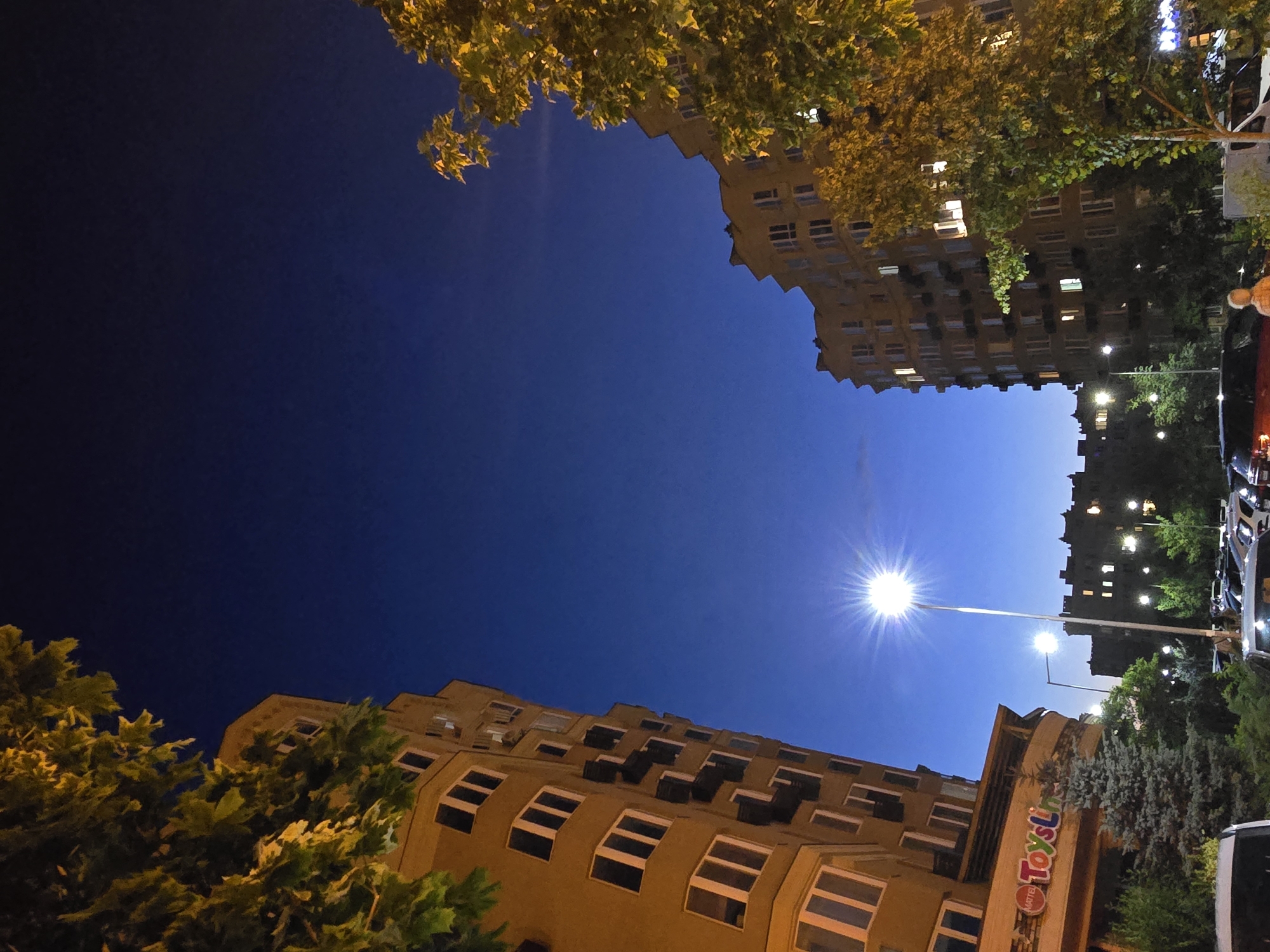 The image captures an upward-looking view of buildings and trees against a deep blue night sky. The scene appears to be an urban or suburban setting, possibly in Sofiivska Borshchahivka, Ukraine, given the provided location context.
In the foreground, lush green trees with yellow-tinged leaves frame the lower portion of the image on the left. Behind the trees, a tan-colored building with numerous windows is visible, some of which are illuminated from within, suggesting occupancy. The building's architecture appears somewhat dated, with a curved facade and recessed window sections.
Further in the distance, another multi-story building is seen, also featuring lit windows and a more solid, darker facade. A bright streetlamp with a starburst effect from its light source dominates the mid-ground, casting light across the scene. Another, less intense light source is visible to the right of the main streetlamp.
On the right side of the image, there is a glimpse of what appears to be a vehicle or bus, with its lights on. Adjacent to the tan building, a sign with brightly colored lettering reads "Toyslim".
The time of day is clearly night, with the deep blue of the sky and artificial lighting being the primary sources of illumination. The weather appears clear, as there are no indications of rain or clouds. The overall mood is one of a quiet evening in a residential or commercial area.