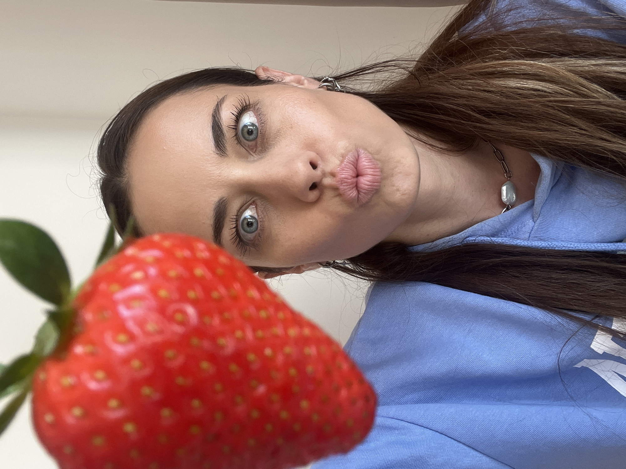 A woman with long brown hair and blue eyes is posing for a selfie with a large strawberry. She is wearing a blue hoodie and a silver necklace. Her face is close to the camera and she is puckering her lips. The strawberry is in the foreground, out of focus. The background is a plain white wall. The image appears to have been taken indoors with natural lighting. There is no contextual information to suggest the location of Bar, Montenegro.
