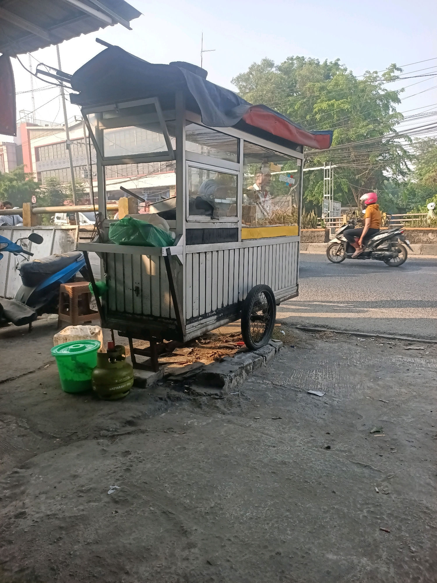 The image depicts a street scene in Gandasari, Indonesia, bathed in the soft light of early morning or late afternoon. A food cart, likely selling fried snacks given the text on the glass display, is positioned prominently on the sidewalk. The cart is a mobile structure, with a white paneled body and a metal frame, topped with a dark fabric cover and a red trim. A single wheel is visible on the right side of the cart. In front of the cart, a green plastic bucket and a green gas cylinder are placed on the ground. To the left of the cart, a blue scooter is parked, partially obscured by the cart.
Further down the street, a person wearing a yellow shirt and a pink helmet is riding a motorcycle. The road is paved and appears to be a public thoroughfare, with power lines strung overhead. Trees with lush green foliage line the background, suggesting a somewhat suburban or semi-urban setting. Buildings are visible in the distance, indicating the presence of more developed areas. The overall impression is one of a bustling street scene, typical of a developing city.