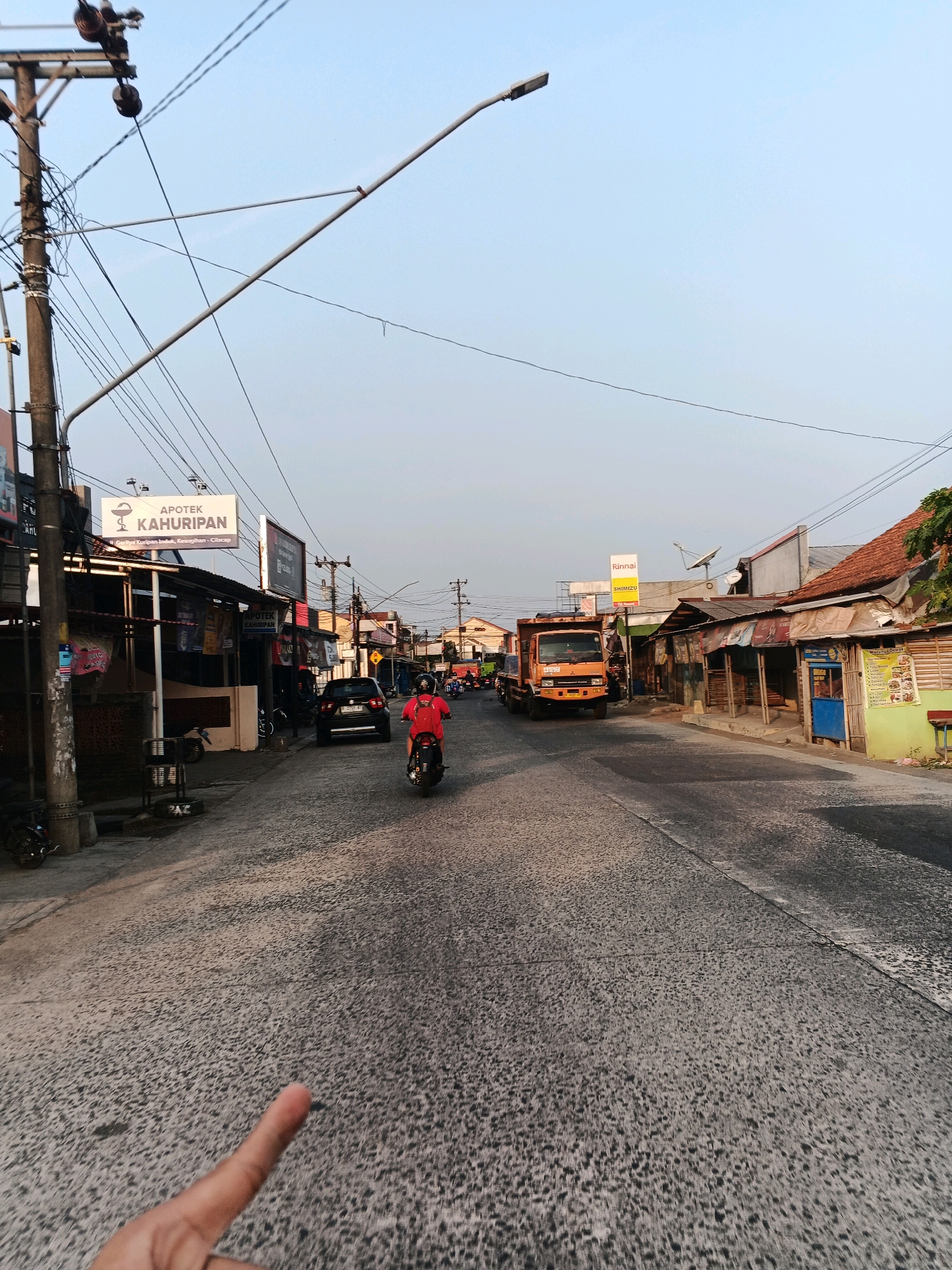 The image depicts a street scene in Cilacap, Indonesia, likely during the late afternoon or early evening due to the warm lighting and long shadows. The street is paved with asphalt and appears to be a moderately busy thoroughfare.
On the left side of the road, there is a prominent utility pole with numerous wires extending across the street. Adjacent to the pole, there are several small shops or stalls with awnings and signage, including one for "APOTEK KAHURIPAN" (Kahuripan Pharmacy). A black car is parked on the left side of the road.
In the center of the image, a person is riding a motorcycle, wearing a red shirt and a helmet. The motorcycle is moving away from the viewer. Further down the street, an orange truck is visible, also moving away.
The right side of the road features a row of buildings, some with corrugated metal roofs and simple facades, suggesting a mix of residential and commercial structures. There are also signs and advertisements on these buildings.
The sky is a clear, pale blue, indicating fair weather. The overall impression is of a typical urban street in Indonesia, with the characteristic mix of infrastructure, businesses, and transportation. The presence of a finger in the foreground suggests the photo was taken from a first-person perspective.