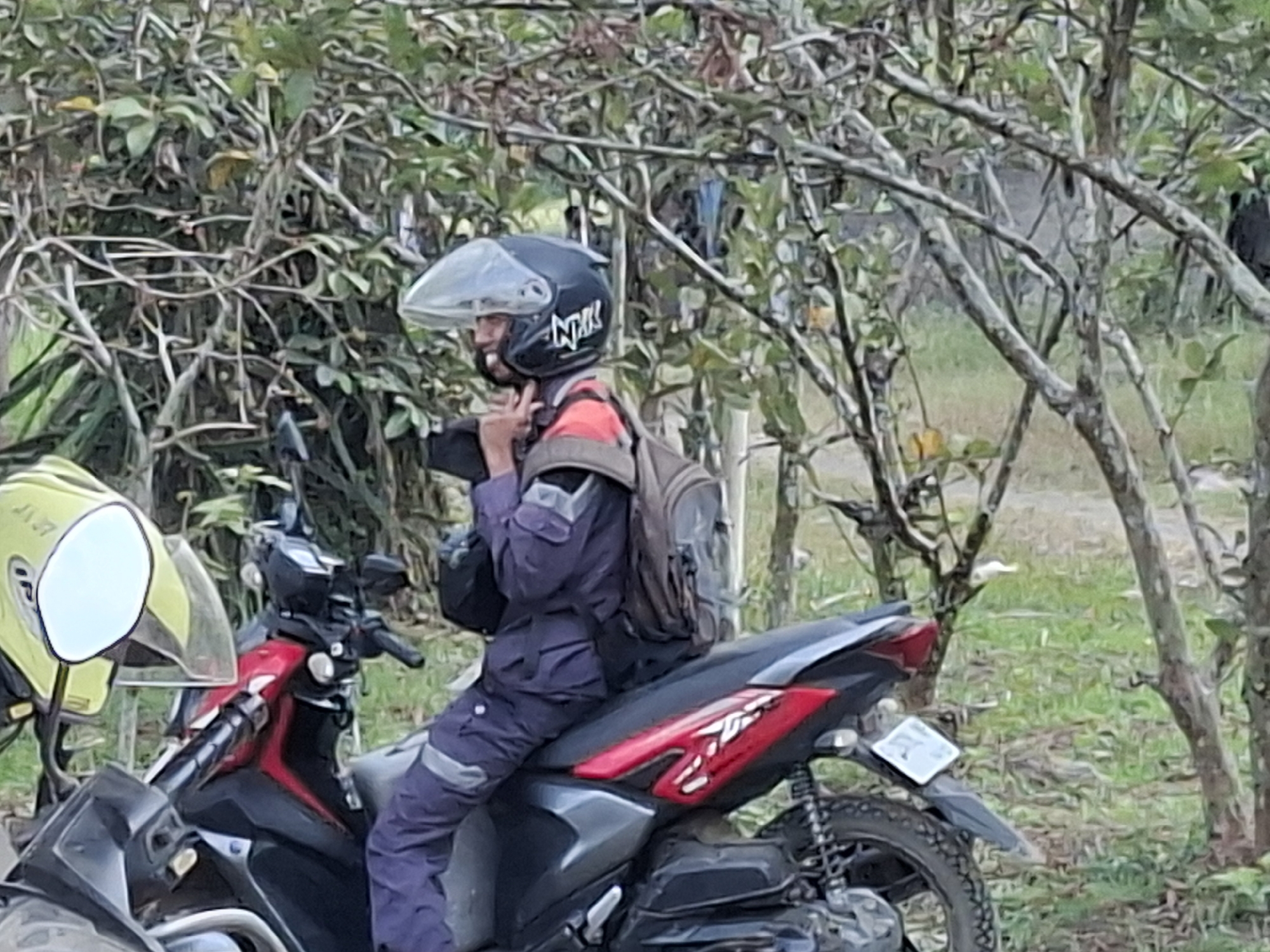 A person wearing a dark blue riding suit and a black helmet with a clear visor is seated on a red and black motorcycle. The person is adjusting their helmet strap. A backpack is visible on their back. A yellow helmet with a white visor is attached to the left side of the motorcycle. The motorcycle is parked on a grassy area with trees and foliage in the background. The scene appears to be in a rural or natural setting.