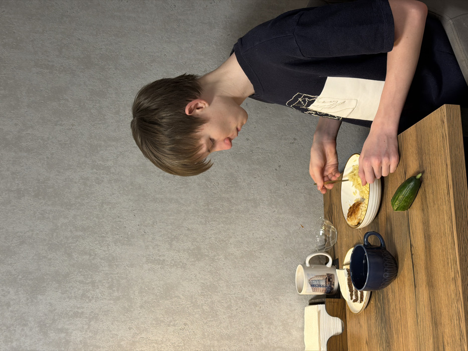 The image, rotated 90 degrees clockwise, depicts a young person, likely male, engaged in eating at a wooden table in an indoor setting.
The main subject is positioned on the right side of the image (when viewed upright). He has short, light brown hair with bangs and is seen in profile, looking down intently at a plate of food. He is wearing a dark blue or black short-sleeved t-shirt with a large white graphic design on the front, which appears to be an abstract outline or a simplified landscape, though specific details are not discernible. His right hand holds a golden fork, actively scooping food from a white, oval-shaped plate that has a thin golden rim. His left hand rests near the plate.
On the white plate, there is a portion of yellow, fluffy food, resembling scrambled eggs or a similar dish, next to a darker, possibly toasted or fried food item.
The wooden surface, likely a table, extends across the lower portion of the image. Arranged on this table are several other items:
* Immediately to the right of the food plate is a whole, dark green cucumber or zucchini.
* Below the food plate, there's a clear, empty glass or small bowl.
* Next to the glass is a white ceramic mug with a colorful graphic print, possibly depicting a building or street scene, but any text or precise details are too small to read clearly.
* A dark blue ceramic mug with a textured surface is positioned next to the white mug.
* Further down on the table, a stack of white paper napkins or folded towels is visible.
The background is a plain, light grey wall with a subtle, rough texture, suggestive of concrete or stucco. The lighting appears to be ambient indoor light. The overall atmosphere suggests a casual mealtime. No explicit time of day or location is identifiable beyond an indoor setting, likely a home. No clearly legible text is visible in the image.