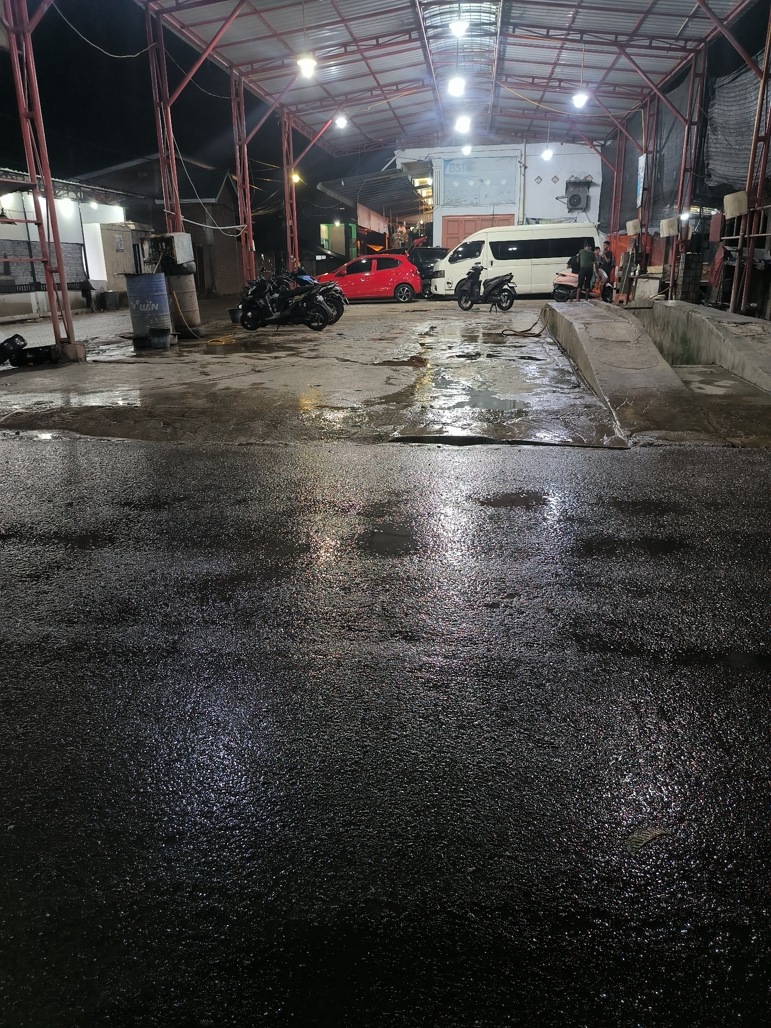 The image depicts a nighttime scene under a covered, open-sided structure, likely a car and motorcycle wash or service station, in Lhokseumawe, Indonesia. The ground is extensively wet and highly reflective, suggesting either recent rainfall or ongoing washing activities.
Under the metal-roofed structure, supported by red painted beams, several vehicles are present. A red hatchback car is parked on the left side, with two dark-colored motorcycles near it. Towards the center-right, a white passenger van is visible, accompanied by at least two other motorcycles. Several people, approximately two to three individuals, are standing and conversing near the white van.
Notable objects include blue barrels, one of which clearly displays the text "H WIN," along with various hoses scattered on the ground. Concrete ramps and drainage systems are visible on the right side of the frame, consistent with vehicle service bays. Artificial lights attached to the roof structure illuminate the scene, creating bright reflections on the wet ground. In the background, other buildings with lit windows are visible, and the letters "bsi" can be seen on a white structure behind the van. The scene conveys a functional, everyday urban environment operating after dark.