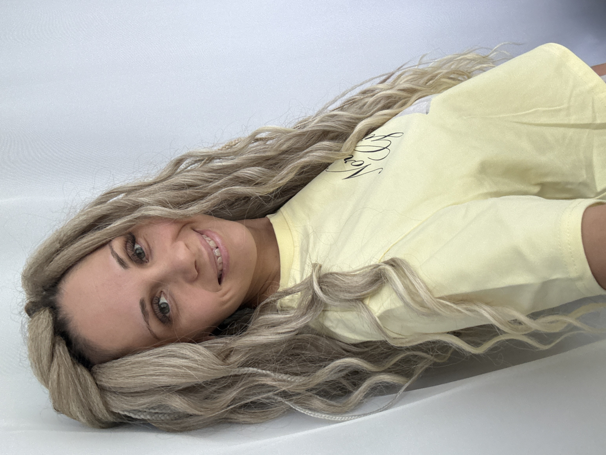 A woman with long, blonde braided hair is lying down on a white surface. She is wearing a light yellow t-shirt with black cursive writing on it. She is smiling and looking at the camera. The background is a solid white color. The image does not contain the location of Rusanovo, Russia.