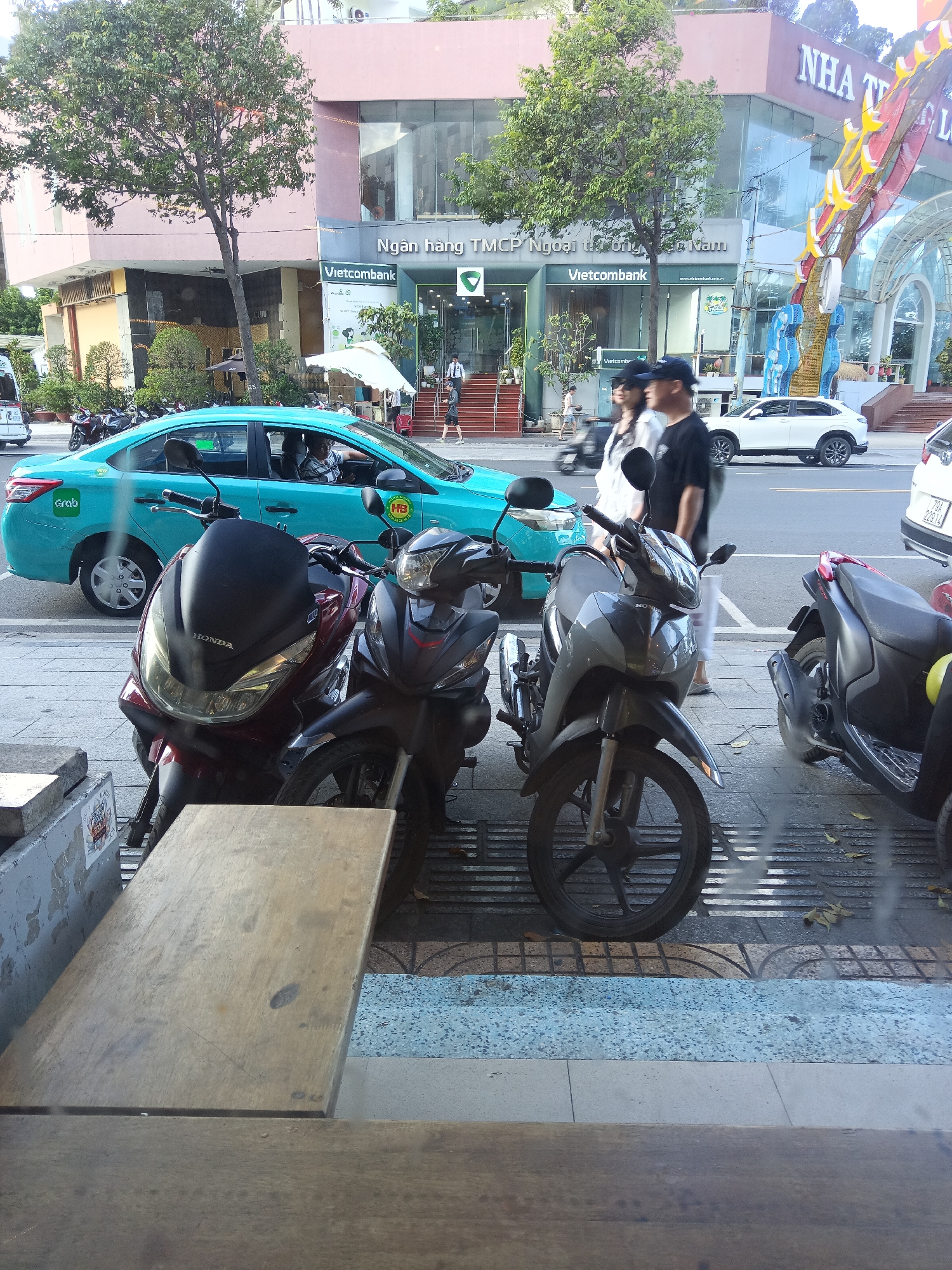 The image shows a street scene in Nha Trang, Vietnam. In the foreground, two motorcycles are parked, a red Honda PCX and a black scooter. Behind them, a teal Grab car is visible, with a driver inside. The car is on the street, and the motorcycles are on the sidewalk. In the background, a building with a Vietcombank branch is present, along with other businesses. Several people are walking on the sidewalk and crossing the street. The scene appears to be a typical urban environment, with traffic, businesses, and pedestrians. The overall atmosphere is one of activity and commerce.