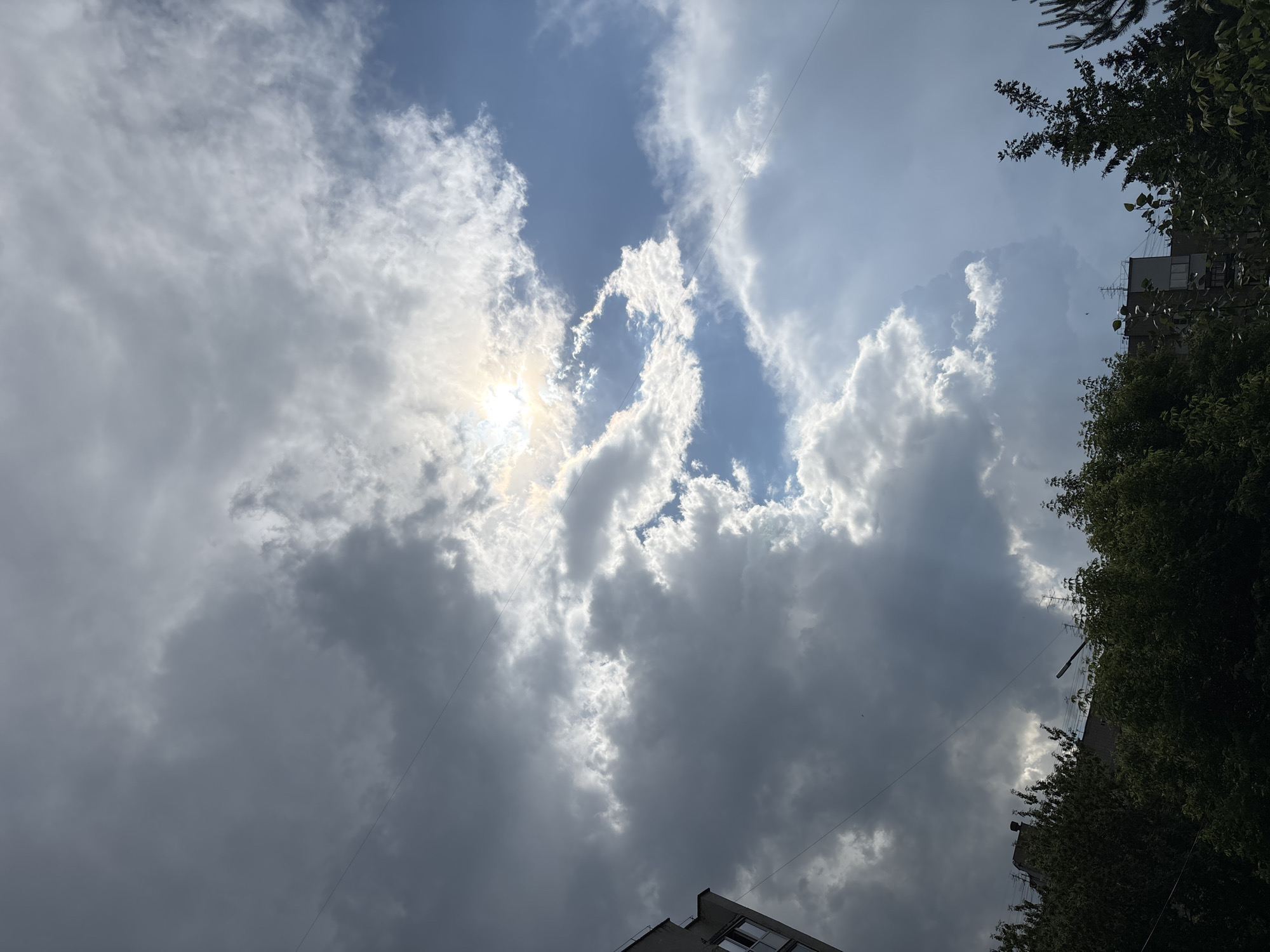 The image captures a dynamic sky over a city, with a mix of dark, heavy clouds and brighter patches where the sun is breaking through. The sun is partially obscured, casting rays of light that illuminate some of the clouds, creating a dramatic effect. The sky transitions from a deep gray to a lighter blue, suggesting a change in weather.
In the foreground, the tops of trees are visible, their dark green foliage contrasting with the bright sky. A portion of a building, likely an apartment block with windows, is also visible on the left edge of the frame. Electrical wires are strung across the sky, a common sight in urban environments. The overall scene is one of a cloudy day with the sun attempting to assert its presence.
The location is identified as Novosibirsk, Russia. This specific urban setting is characterized by its residential buildings and the presence of trees, indicating a part of the city with greenery. The weather conditions suggest a temperate climate, possibly late spring, summer, or early autumn, given the presence of lush green trees. No people or specific activities are depicted.