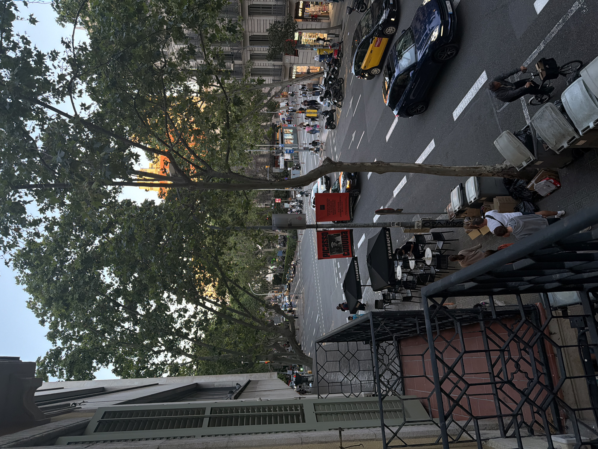 The image depicts a street scene in Barcelona, Spain, observed from an elevated viewpoint. It is daylight. In the foreground, there is a metal balcony railing. Adjacent to the railing, some waste containers are visible along the edge of the sidewalk. A person is lying on the sidewalk. Further along the sidewalk, an outdoor Starbucks is set up with umbrellas, tables, and chairs. A street extends into the distance, with a banner advertising "Picasso Convida." Pedestrians populate the street, and there are various vehicles, including cars, a taxi, and motorcycles. Buildings line both sides of the street. Trees with dense foliage are interspersed between the buildings and along the street.