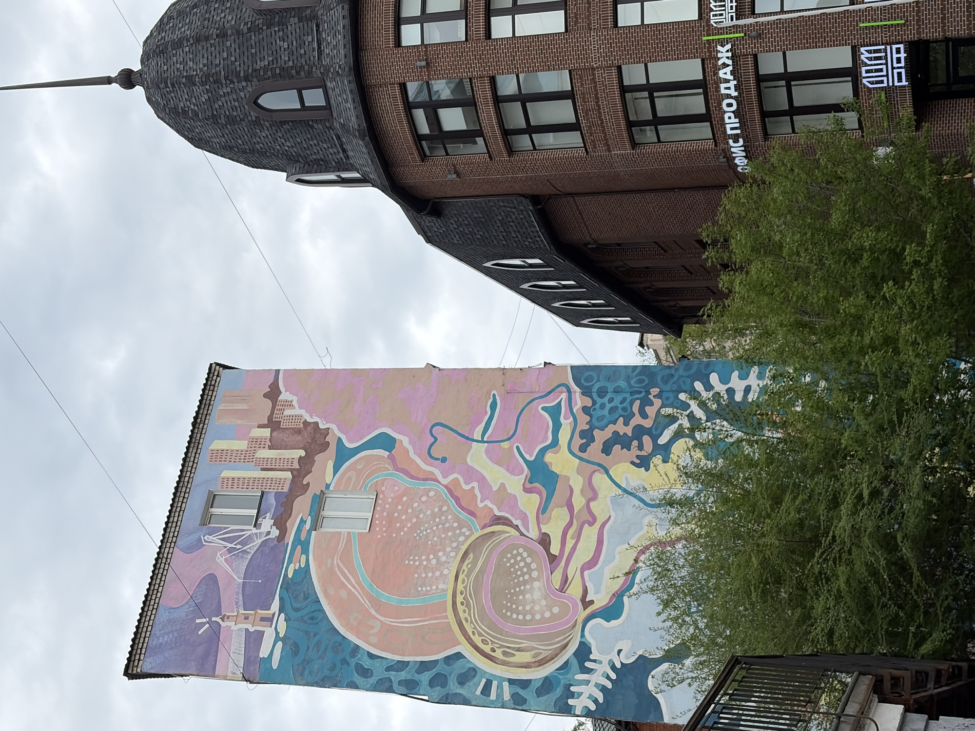 The image shows an upward view of two buildings in Vladivostok, Russia, under a cloudy sky. On the left, a building is adorned with a large mural featuring a fantastical landscape and organic shapes, including a large, peach-colored circular form, possibly representing a mollusk or sea creature. Architectural elements of Vladivostok are incorporated into the mural's design.
To the right, a taller brick building features a dark, conical roof with a spire. Rectangular windows are regularly spaced across the facade. A sign with vertical lettering reading "офис продаж" ("sales office") is visible, along with other illuminated vertical signs. A tree with green foliage partially obscures the building. A black metal structure is visible in the foreground near the mural. Overhead electrical wires stretch across the sky.
