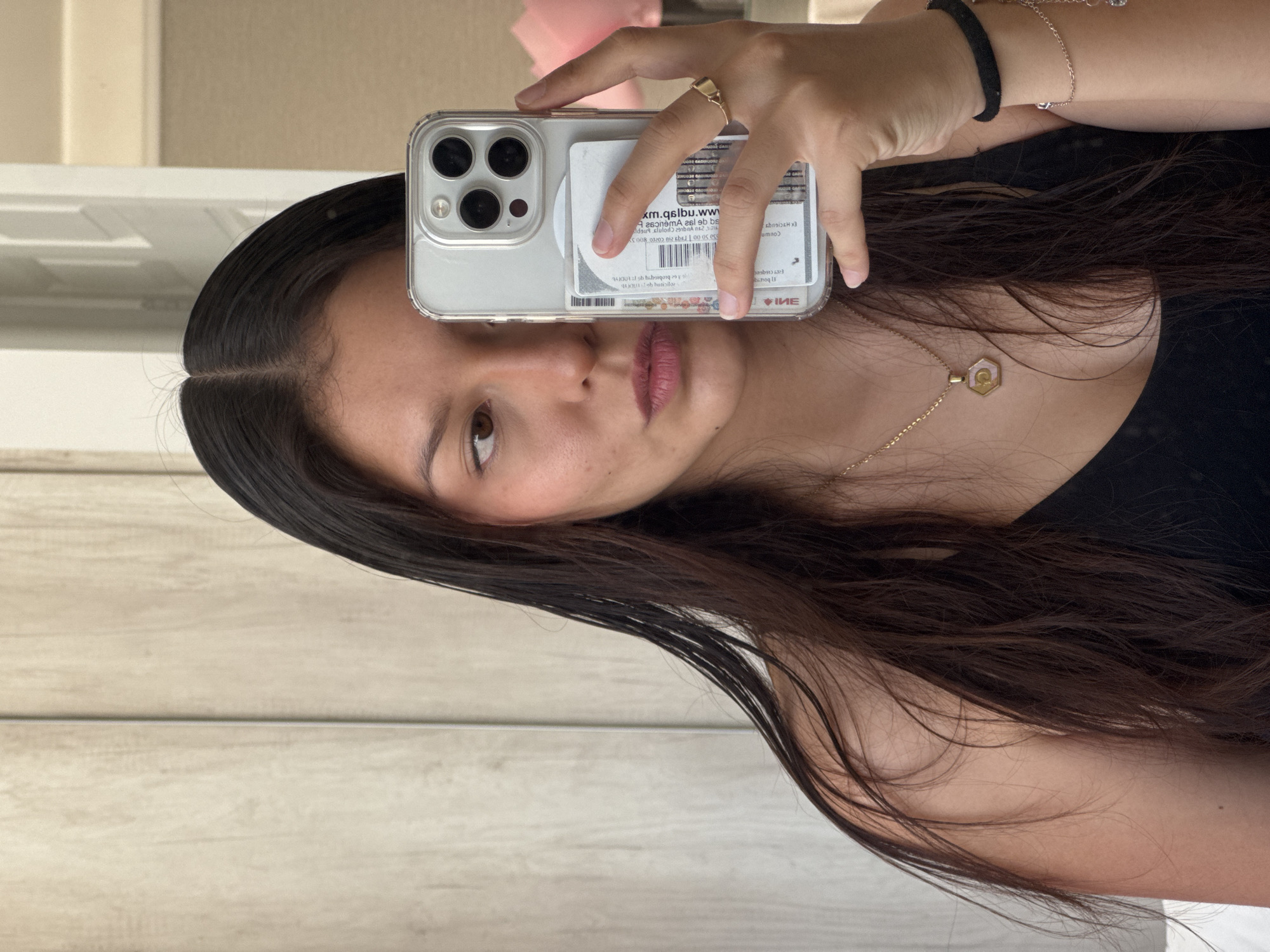 A mirror selfie features a young woman, captured in a portrait orientation within a horizontally taken photograph, implying the image itself is rotated 90 degrees counter-clockwise. She has long, dark hair parted down the middle, dark eyes, and wears pinkish lipstick, maintaining a neutral expression. She is dressed in a black top or dress.
Her right hand holds an iPhone with a clear case horizontally in front of her face, with the phone's triple camera lenses clearly visible. Behind the phone, several cards are stored within the transparent case. The text on these cards appears reversed due to the mirror reflection. Partially discernible reversed text includes "www.ubsq.mx" (likely a website), and a sequence like "00.00 leba zin 202300 800 SK" (which strongly suggests numbers and a date from an identification card or document). Other reversed fragments like "НИЕ" and "umno" are also visible. Given the location context of San Francisco Ocotlán, Mexico, these documents are likely Mexican.
Her left arm is visible, adorned with a black hair tie and a silver-colored bracelet. A gold-toned ring is on a finger of her right hand, and she wears a delicate gold chain necklace with a hexagonal pendant that features a circular symbol. The background is simple and light-colored, appearing to be an indoor setting with either a plain wall or light wooden paneling, under what appears to be daytime lighting. The overall image captures a casual self-portrait.