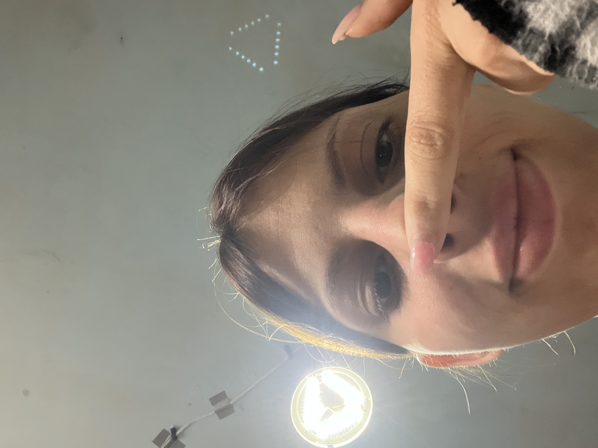 A close-up, skewed perspective image features a woman indoors, holding up her index finger towards the camera. Her face is prominently visible, including her eyes, nose, and lips. A lighting fixture is visible in the lower portion of the frame, possibly mounted on the ceiling. The background is a plain, light-colored surface. The location is not discernible.