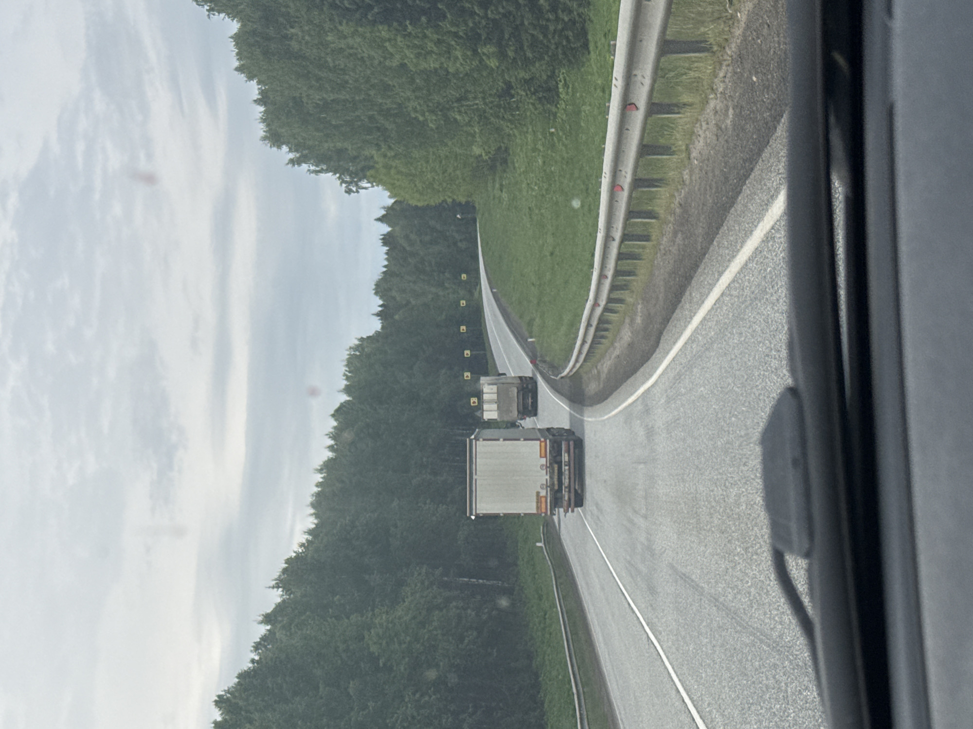 The image, taken from inside a vehicle, captures a view of a two-lane asphalt road curving to the right through a forested area on an overcast day. The scene shows the perspective of someone driving on the road, with part of the vehicle's interior visible on the right side of the frame.
In the foreground and midground, two large trucks with white trailers are visible, traveling in the same direction as the viewer's vehicle. The road surface appears damp, suggesting recent rain or drizzle. A metal guardrail with red reflectors lines the right side of the road. Both sides of the road are bordered by dense green coniferous or mixed forest, extending into the distance.
Along the left side of the road, near the tree line, several small yellow square markers or signs are visible, spaced out. The sky above is largely covered by light grey clouds, indicating an overcast condition. The overall impression is of a journey through a rural or wilderness area. No specific text is legible in the image. The location context for this scene is Verkhnyaya Talitsa, Russia.