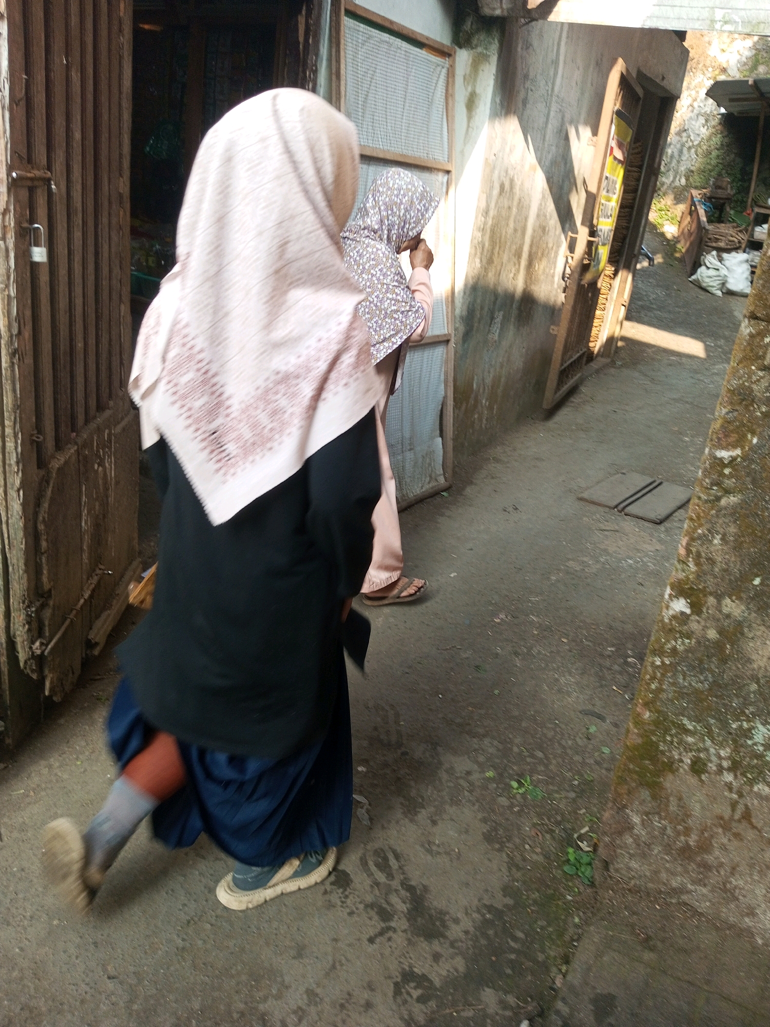 This is a medium-shot, eye-level, outdoor photograph taken in Kalibeber, Indonesia, showing two individuals walking down a narrow, somewhat cluttered alleyway. The primary subject, on the left, is a person from the back, wearing a light pink hijab with a subtle pattern and a dark long-sleeved shirt over a dark blue skirt. This individual is in motion, with their left leg extended forward, appearing to be walking briskly or running. They are wearing light grey socks and dark sneakers with thick, off-white soles. The right subject, further down the alley, is partially visible from the side, wearing a patterned hijab and a light-colored garment, also in motion.
The environment is characterized by weathered wooden structures on the left, including a door with a padlock. The wall on the right is rough and textured, with a mossy appearance in places. The ground is a rough, dusty concrete or asphalt surface with some scattered debris and small patches of green vegetation. The lighting suggests it is daytime, with shadows cast by the structures. In the background, other structures, possibly stalls or buildings, and some stacked materials are visible, hinting at a market or a densely populated area.