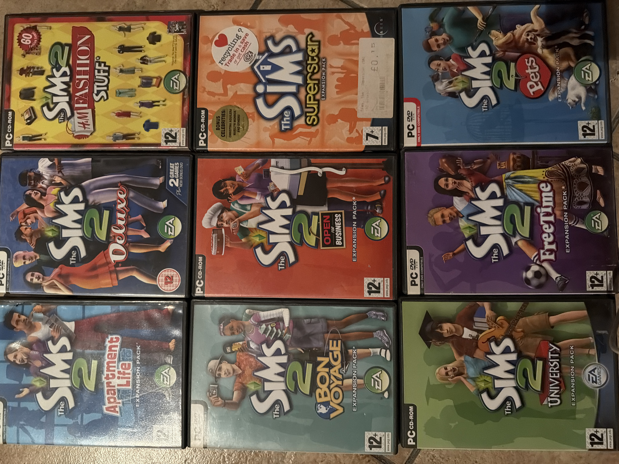 The image displays a collection of PC game cases for "The Sims 2" expansion packs, arranged in a grid on a textured surface. The games are identified as "Apartment Life," "Deluxe," "H.M. Fashion Stuff," "Bon Voyage," "Open for Business," "Superstar," "University," "FreeTime," and "Pets." Several of the cases have PEGI age rating symbols (12+ or 7+) and EA Games logos. One case, "H.M. Fashion Stuff," features a "60 fashion items!" badge. The "Superstar" case has a sticker that reads "Recycling? Trade in & save or get cash" and a price tag of "£0.15." The overall condition of the cases appears to be used, with some minor scuffs and wear visible. There is no indication of the city of Sharm El Sheikh, Egypt, in the image.