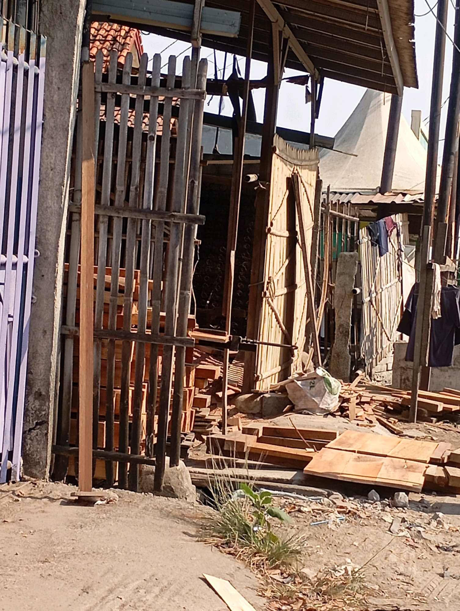 In Babelan, Indonesia, an outdoor scene depicts a construction or workshop area. A light purple, vertical slat fence is visible on the left. Adjacent to it is a grey concrete pillar. In the center, a rustic gate constructed of vertical bamboo poles bound by horizontal wooden planks stands open. Behind this gate, stacks of rectangular, light brown wooden planks are organized. Further back, a wooden structure with a corrugated roof is partially visible, with clothing hanging from lines. A conical, light-colored structure, possibly a kiln or chimney, rises in the background. The ground is a mixture of dirt and gravel, with debris, scattered wooden offcuts, and a white plastic bag containing some material. The overall impression is one of a working site, possibly for woodworking or building.