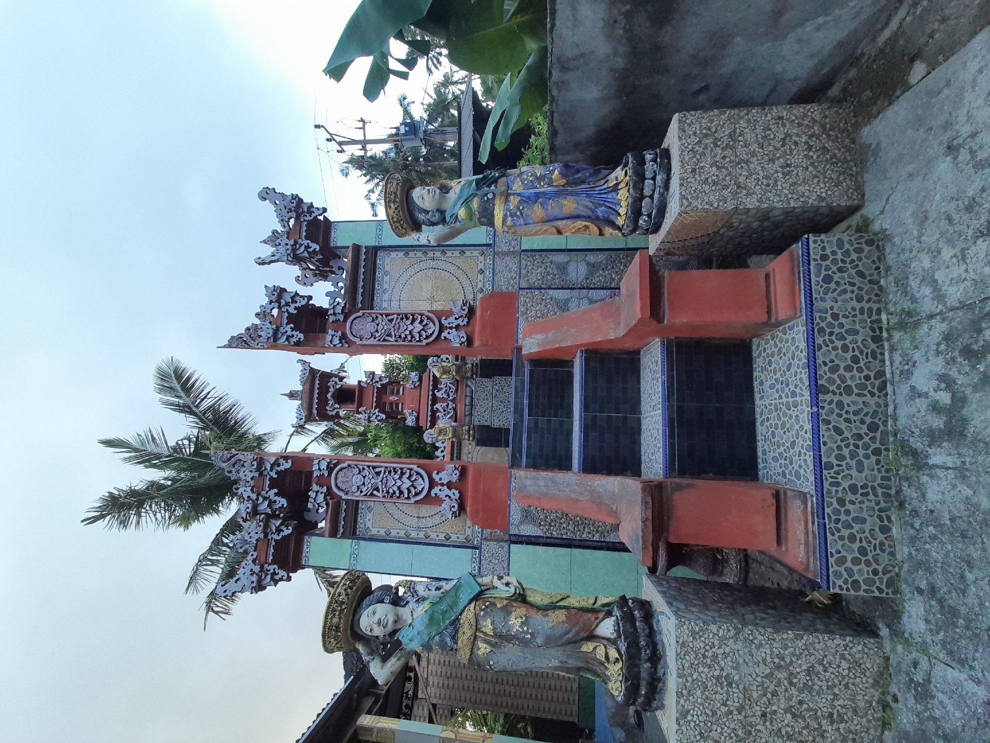 The image displays an outdoor scene, rotated 90 degrees counter-clockwise, capturing an ornate entrance flanked by two statues. When viewed upright, the central feature is a traditional, multi-tiered gateway, reddish-brown in color, adorned with intricate white and gold carvings. The gateway's design suggests a Balinese or Southeast Asian architectural style.
Two female statues guard the entrance, one on each side. Both are depicted wearing elaborate headwear and traditional, colorful attire with prominent blue, gold, green, and dark brown accents. The statue on the left (when viewed upright) has her left arm raised, while the one on the right has her right arm resting. They stand on textured bases, which are part of a larger tiled area.
The ground and walls around the entrance feature various decorative tiles: some are light blue with circular patterns, others are textured with small grey and black stones, and black and blue tiles form steps. In the background, palm trees and other green foliage are visible, along with a utility pole and power lines. The sky is bright and clear, suggesting daytime. No discernible activity is taking place, and there is no visible text.