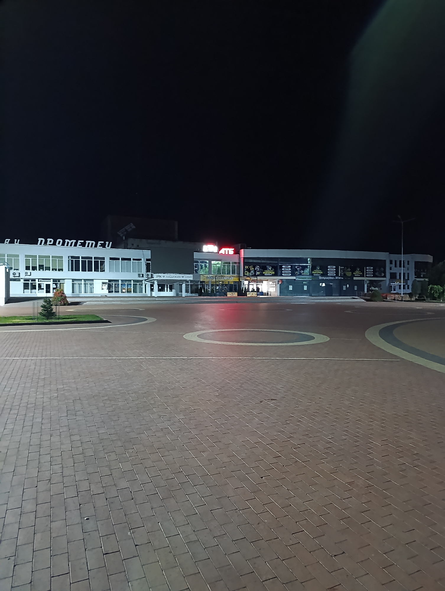 An exterior, eye-level, nighttime photograph captures the facade of a modern, multi-story commercial building at the Prometey shopping and entertainment center in Ladyzhyn, Ukraine. The building is illuminated by artificial lights, highlighting its clean white and dark-gray architectural lines. The entrance to the shopping center is visible, with large glass windows displaying various shops and displays. A small evergreen tree is planted in front of the building on the left side. The foreground of the image is a wide, paved plaza with a distinctive pattern of reddish-brown interlocking pavers, accented by circular inlays of gray pavers. The sky above is dark and devoid of stars. The scene is quiet and appears to be free of people.