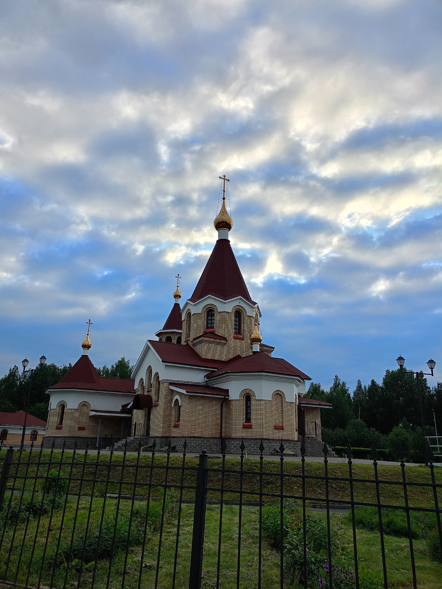 A wide-angle, eye-level shot captures an Orthodox church with a yellow brick facade and a red roof under a cloudy, blue sky. The church features multiple domes, some with gold crosses. The building is partially obscured by a black metal fence in the foreground, behind which is a grassy area with some bushes and small trees. In the background, a dense line of dark green trees extends across the horizon. Two lampposts are visible on the right side of the image. The overall lighting suggests either early morning or late evening.