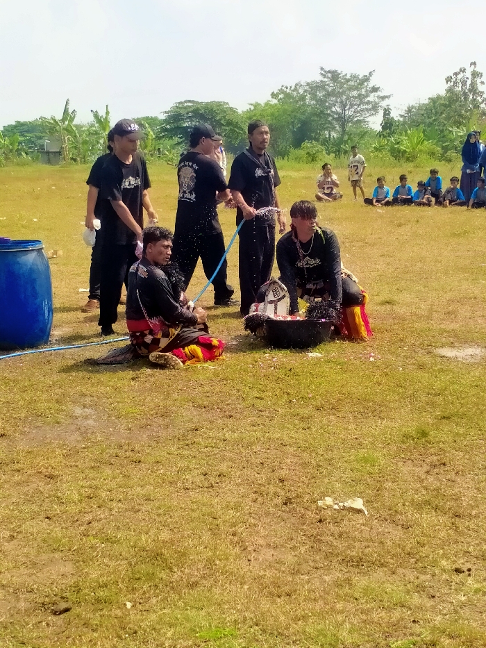 This image depicts an outdoor event on a dry, patchy grass field in Celep, Indonesia, under a clear sky. In the foreground, five adult males, all wearing black shirts and trousers, are involved in a water-based activity. Two men are positioned on the ground; one kneels on the left, partially covered by red and yellow patterned fabric over his black trousers, looking right. The other crouches on the right, similarly adorned, facing forward. Both are being sprayed with water from blue hoses held by two standing men behind them, who are also visibly wet. A third standing man on the far left holds a white cloth. Between the two seated men is a black basin containing water and white floral material. A large blue plastic barrel is on the far left. In the mid-ground, approximately ten younger individuals, primarily males in blue shirts, sit on the grass observing. Further back, a person in a light blue hijab is visible on the right. The background features trees, green foliage, and a distant structure on the far left. The ground immediately surrounding the central activity is wet and muddy.
