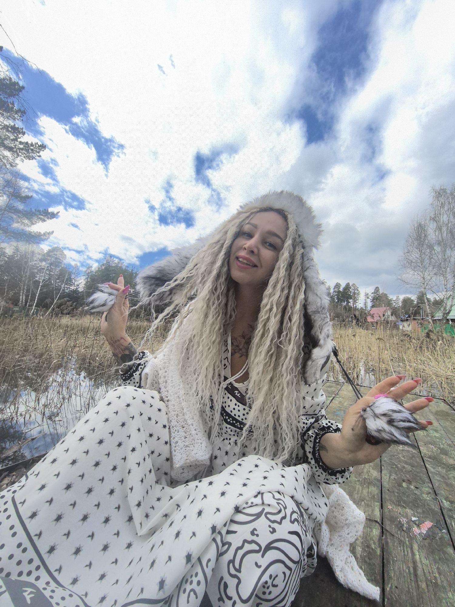A woman with long, curly blonde hair and wearing a white and black patterned outfit with a fur-trimmed hood is seated on a wooden surface near water. She is holding small, fluffy objects in each hand, showing them off to the camera. The background includes a marshy area with tall reeds and trees, and some buildings can be seen in the distance. The sky is cloudy. The photo appears to be taken in a natural setting, possibly near a body of water, in the city of Noviy, Russia.