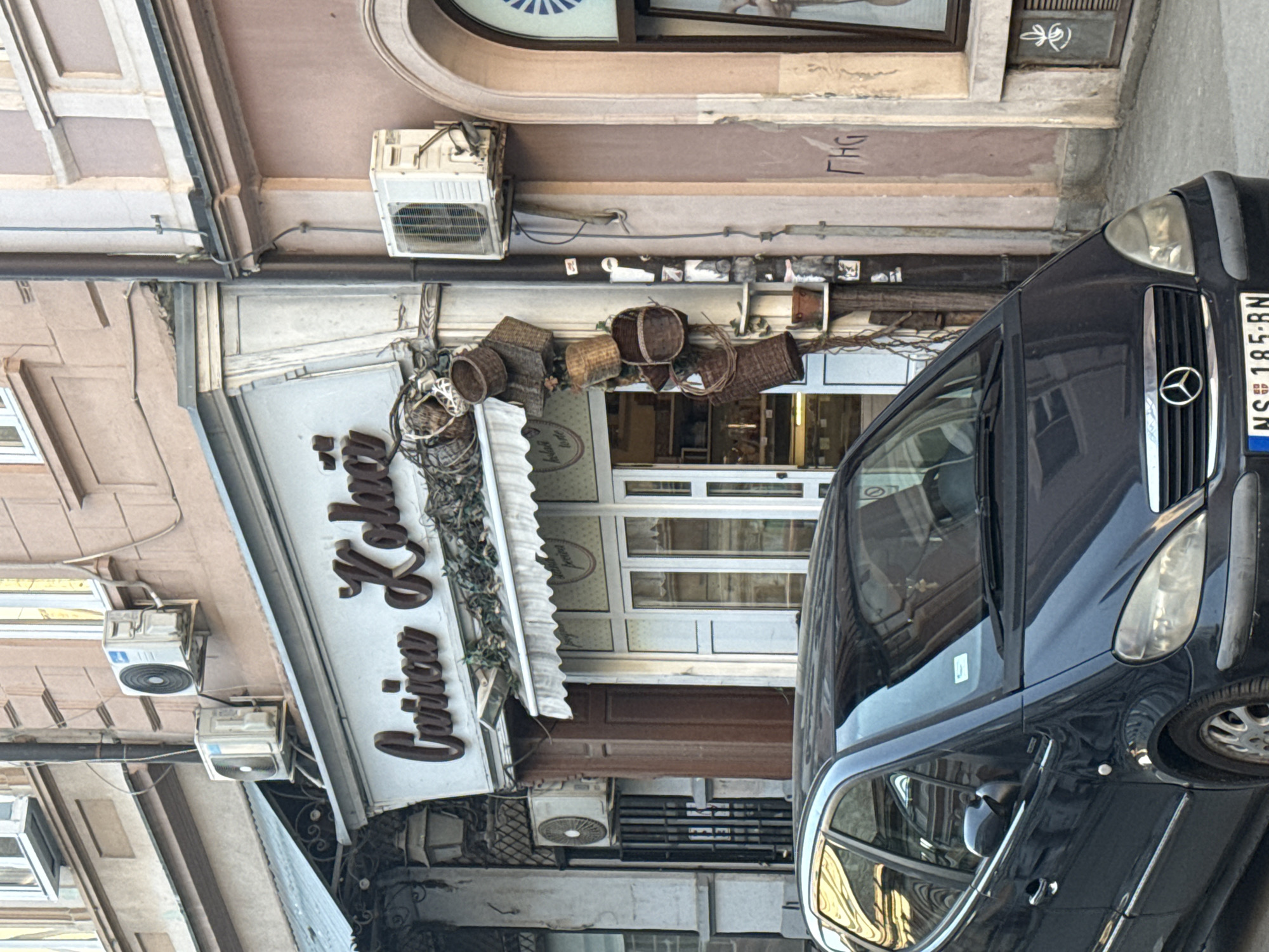 Here is a forensic description of the image:
The image captures a street scene with a dark-colored Mercedes van parked in front of a shop named "Carica Lellian". The shop's facade is white with brown ornate lettering. Several woven baskets decorate the space above the shop's entrance. Multiple air conditioning units are visible on the building's exterior, both above and below the shop's signage. The street appears to be in a city, evidenced by the architecture and the parked vehicle. The license plate of the car is "NS 185-BN". The overall impression is that of an urban street scene with a combination of commercial and residential elements.