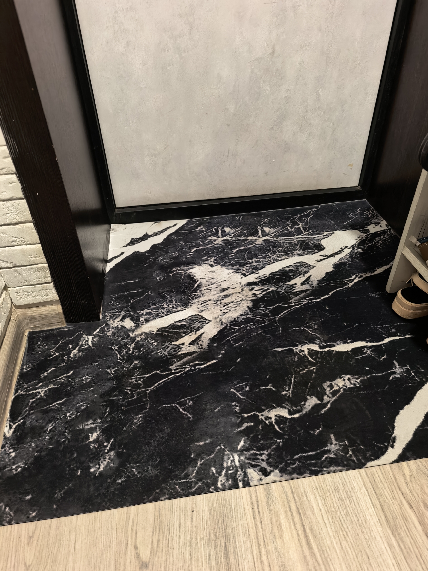 The image shows a close-up, high-angle shot of a black and white marble-patterned rug placed on a light-colored wooden floor. The rug has a striking, vein-like pattern of white against black, resembling natural marble. The rug appears to be in an entryway or hallway, as indicated by the dark wooden door frame and a light-colored wall with a brick pattern on the left side of the image. Above the rug, a light-colored wall with a textured surface, possibly tile or concrete, is visible. The lighting in the scene is somewhat dim, suggesting it might be indoors with artificial lighting, or it's a time of day with low natural light. There is a hint of a shoe rack or shelf on the right side of the image, with a pair of beige sandals visible on it. The scene is static, with no activity taking place. The focus is on the textures and patterns of the rug and the surrounding materials. No text is visible in the image. The setting appears to be a residential interior in Sterlitamak, Russia.