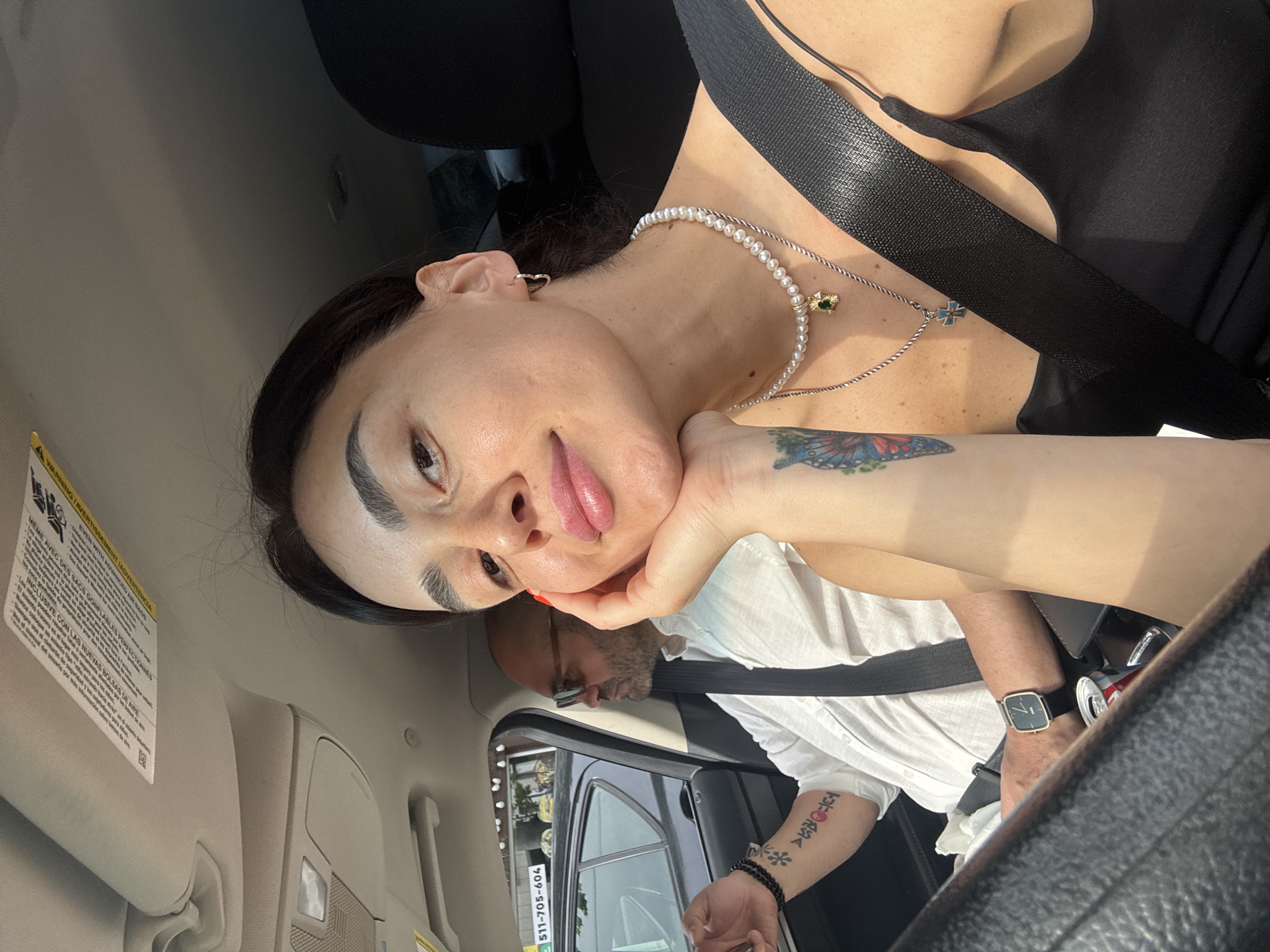 The image shows a woman in the passenger seat of a car, taking a selfie. She has dark hair, is wearing a black tank top, a pearl necklace with a green pendant, and a silver necklace with a blue cross pendant. Her left arm, visible on the left side of the image, has tattoos and is resting on her chin. A man is visible in the driver's seat of the car, wearing a white shirt and glasses. He has a watch on his left wrist. The car appears to be on a street in Tbilisi, Georgia, with buildings visible through the car window. The weather appears to be sunny.