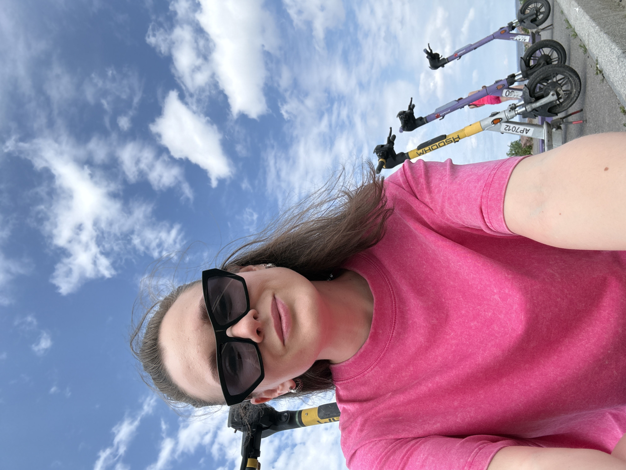 A young woman wearing sunglasses and a bright pink t-shirt takes a selfie outdoors, with a line of electric scooters visible behind her. The sky is blue with scattered white clouds, suggesting a clear day. The woman has her hair pulled back, and is smiling slightly. The scooters are parked side-by-side, with some bearing yellow and purple accents and visible branding. The environment appears to be an urban setting, possibly a waterfront or park area, given the distant hint of water and greenery. The overall scene is casual and summery.