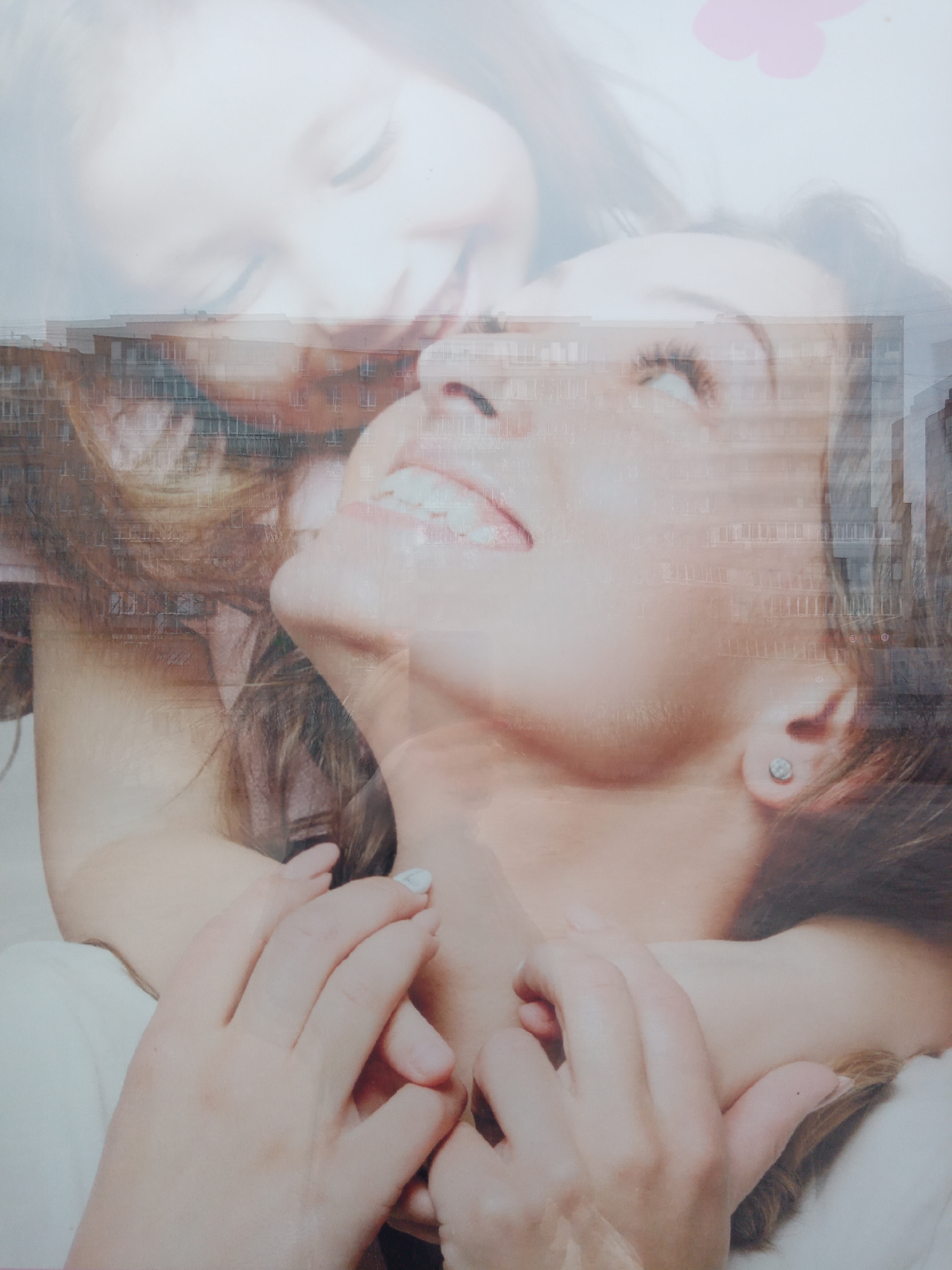 The image appears to be a multiple exposure photograph featuring a woman in the foreground with a transparent overlay of a child's face above her. A building, possibly an apartment complex, is also faintly superimposed onto the woman's face and shoulders. The woman is looking upward with a slight smile, while the child's face is blurred and indistinct. The child's hands are wrapped around the woman's neck. The city and location cannot be determined.