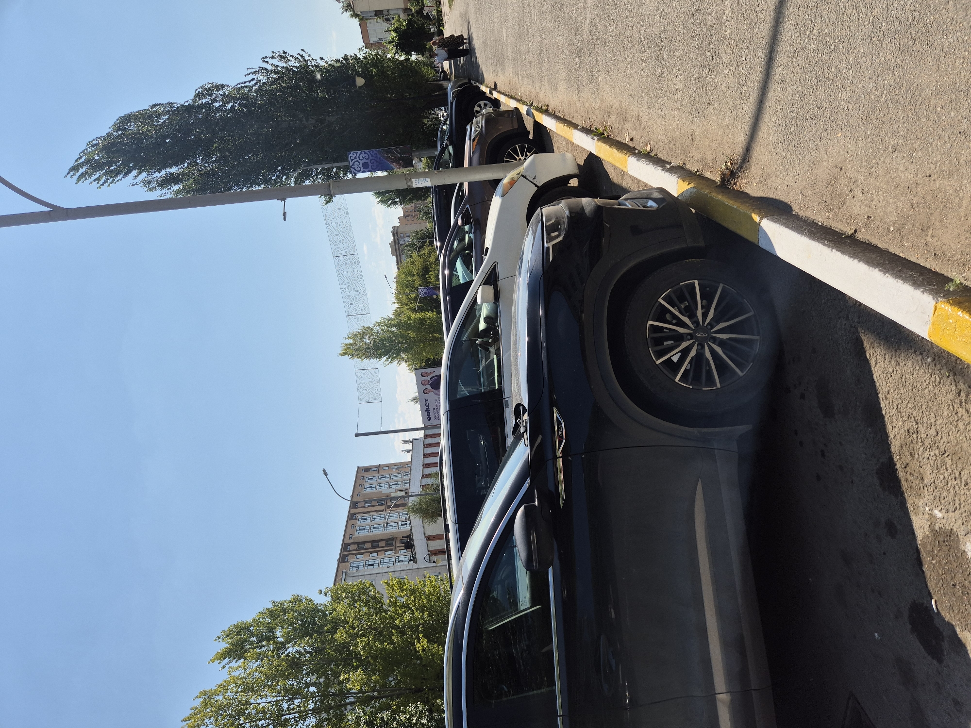 The image shows a street in Kokshetau, Kazakhstan, with several cars parked along the side of the road. The cars are lined up next to a curb, which is painted with alternating yellow and white stripes. In the background, there are buildings and trees, and the sky is blue. The sun is shining, casting shadows on the road. A pedestrian is walking on the sidewalk in the distance. The scene appears to be a typical urban environment on a sunny day.