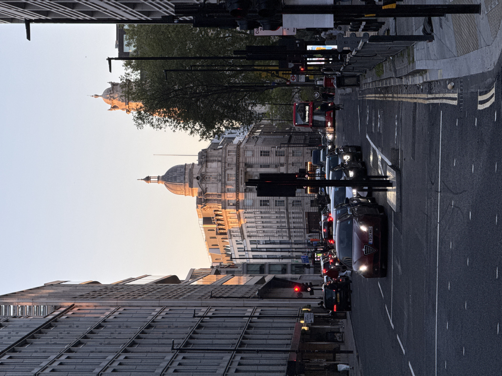 The image shows a street in London, United Kingdom, with buildings on either side and traffic in the road. The buildings are a mix of modern and classical architecture, with one featuring a dome. Traffic includes cars, a red double-decker bus, and motorcycles. The street is lined with lampposts and traffic lights. The sky is light, suggesting either early morning or late afternoon. The road has lane markings and appears to be wet.