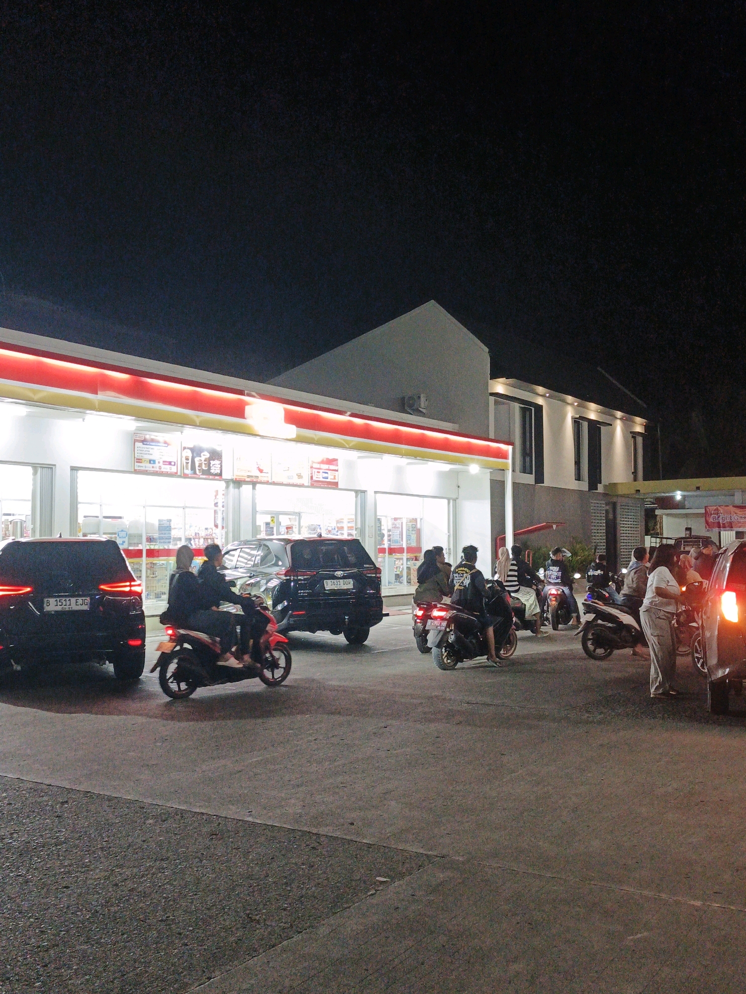 A night scene at a brightly lit convenience store in Pangandaran, Indonesia, depicts a lively atmosphere with several motorcycles and cars present. The store, with its red and yellow trim and illuminated interior displaying products, is a central feature. Various individuals are seen on or around the motorcycles, some appearing to be refueling or interacting. One black SUV with license plate B 1511 EJG is parked on the left, and another black SUV with license plate B 3633 DDU is to the right, both illuminated by their brake lights. Several people are gathered near the motorcycles in front of the store.