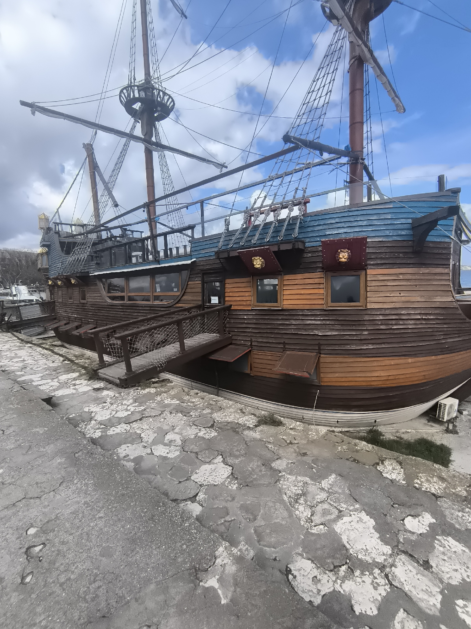 The image depicts a stationary wooden ship designed to resemble a pirate ship, located on a cracked, concrete dock. The hull is brown, with a blue upper section and two closed window shutters. The ship has multiple masts, rigging, and a crow's nest. It is connected to the dock by a wooden gangway with rope netting on the sides. There is an air conditioning unit beneath the ship near the stern. The sky is partly cloudy and blue. The image shows a floating restaurant built like a pirate ship that's located in Varna, Bulgaria.