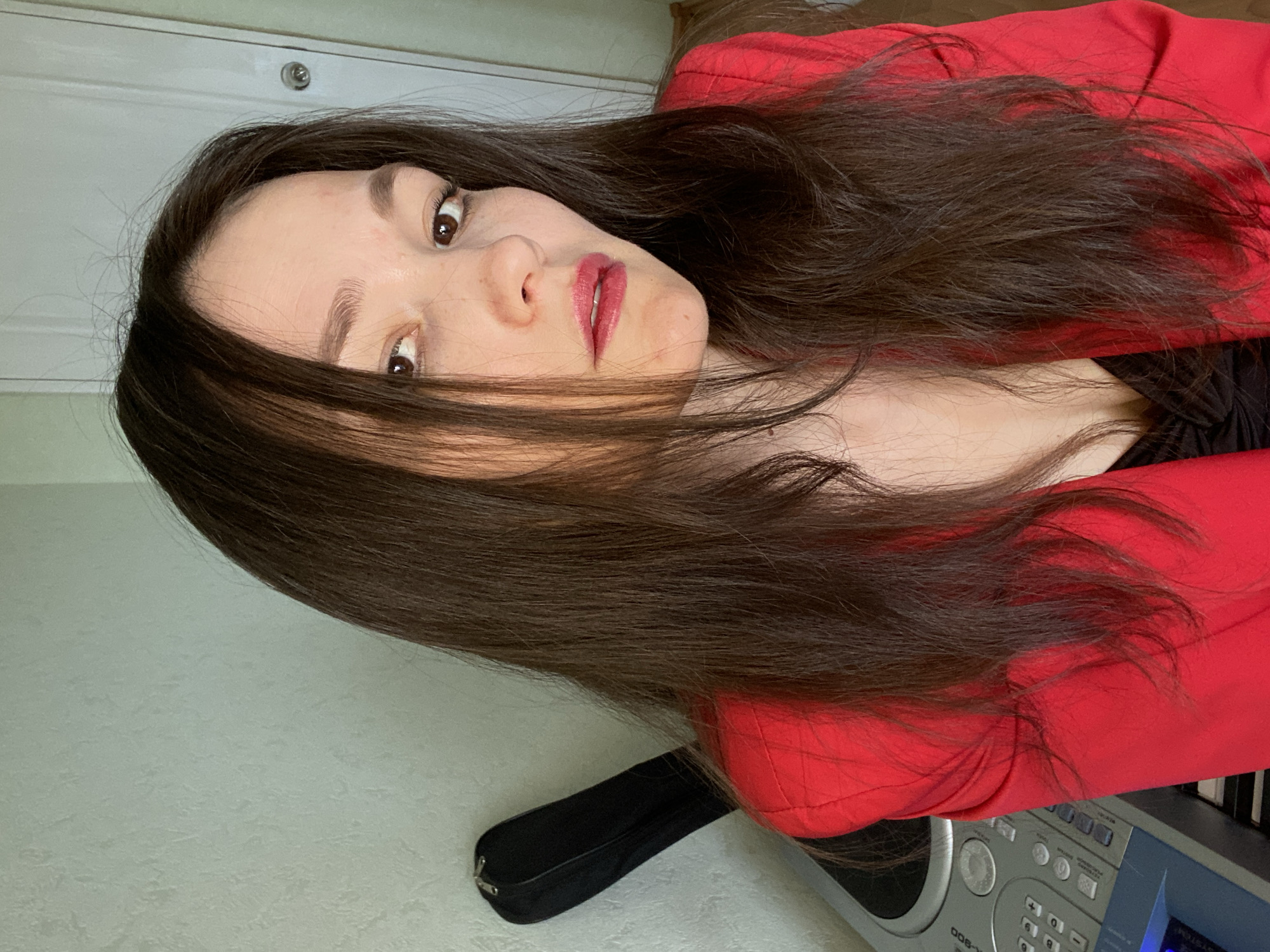 A woman with long, dark brown hair is shown in a close-up, waist-up shot. She is wearing a red blazer over a black top and has makeup on her lips. Her head is tilted slightly to the right, and she is looking directly at the camera. In the background, a light-colored wall with a textured surface is visible, as is a portion of a door with a round doorknob. To the left of the woman, a black case and part of a silver electronic keyboard are visible. The scene appears to be indoors, possibly in a home or studio setting. The image was taken in the city of Chaikovsky, Russia.